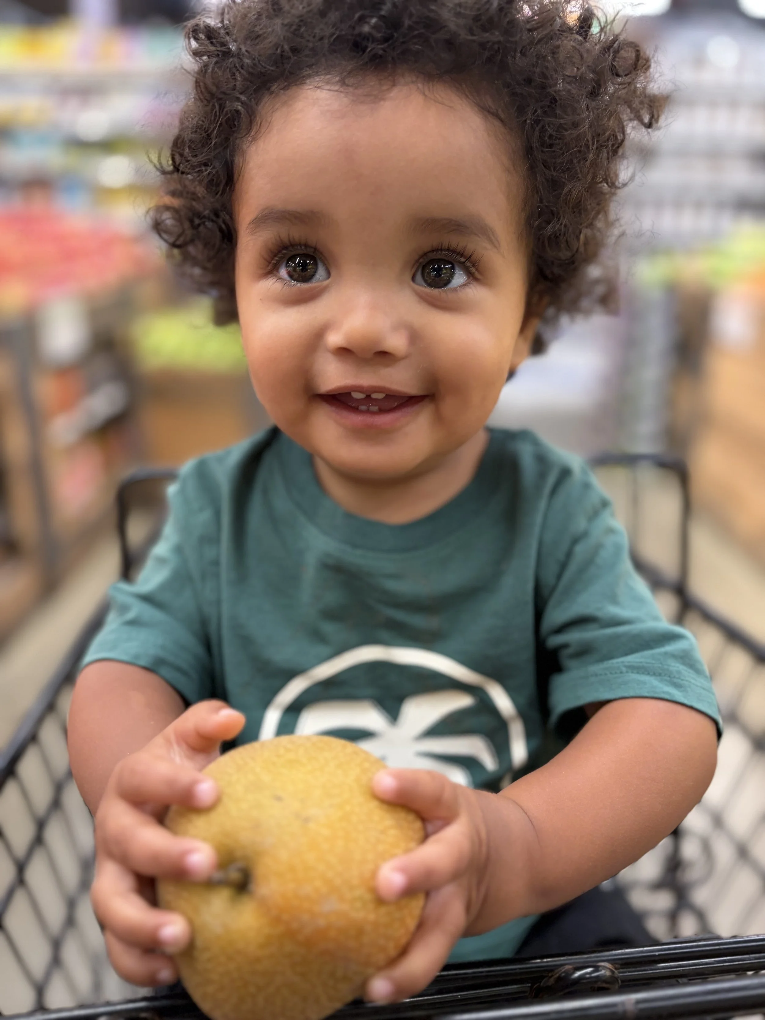 A young child with curly hair and big eyes, sitting in a shopping cart, holding a yellow fruit, possibly a lemon, with an adult's hand supporting it.