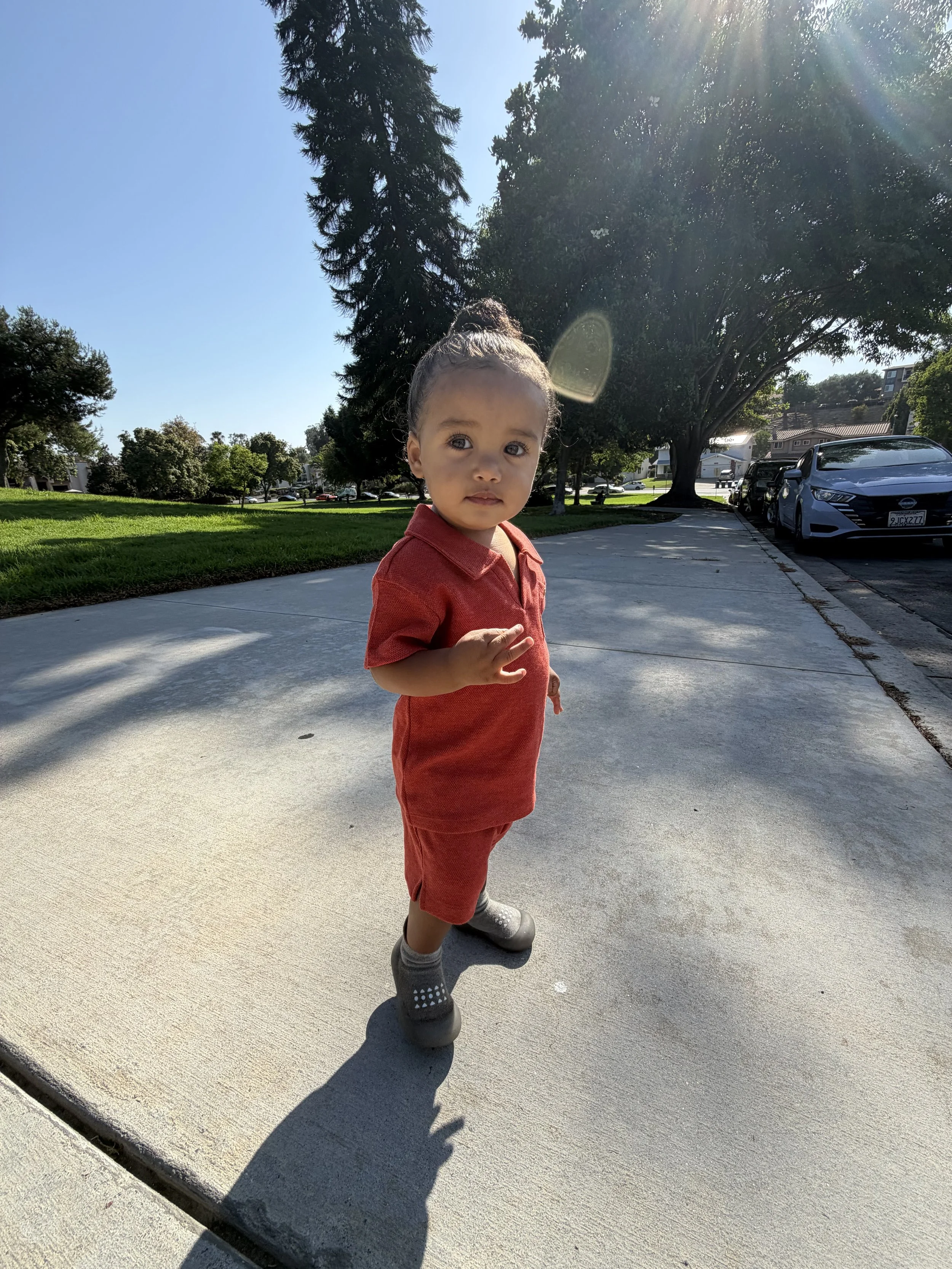 A young child with a top knot hairstyle and a calm expression, wearing a red outfit and gray shoes, standing on a concrete sidewalk in a sunny park with trees and parked cars in the background.