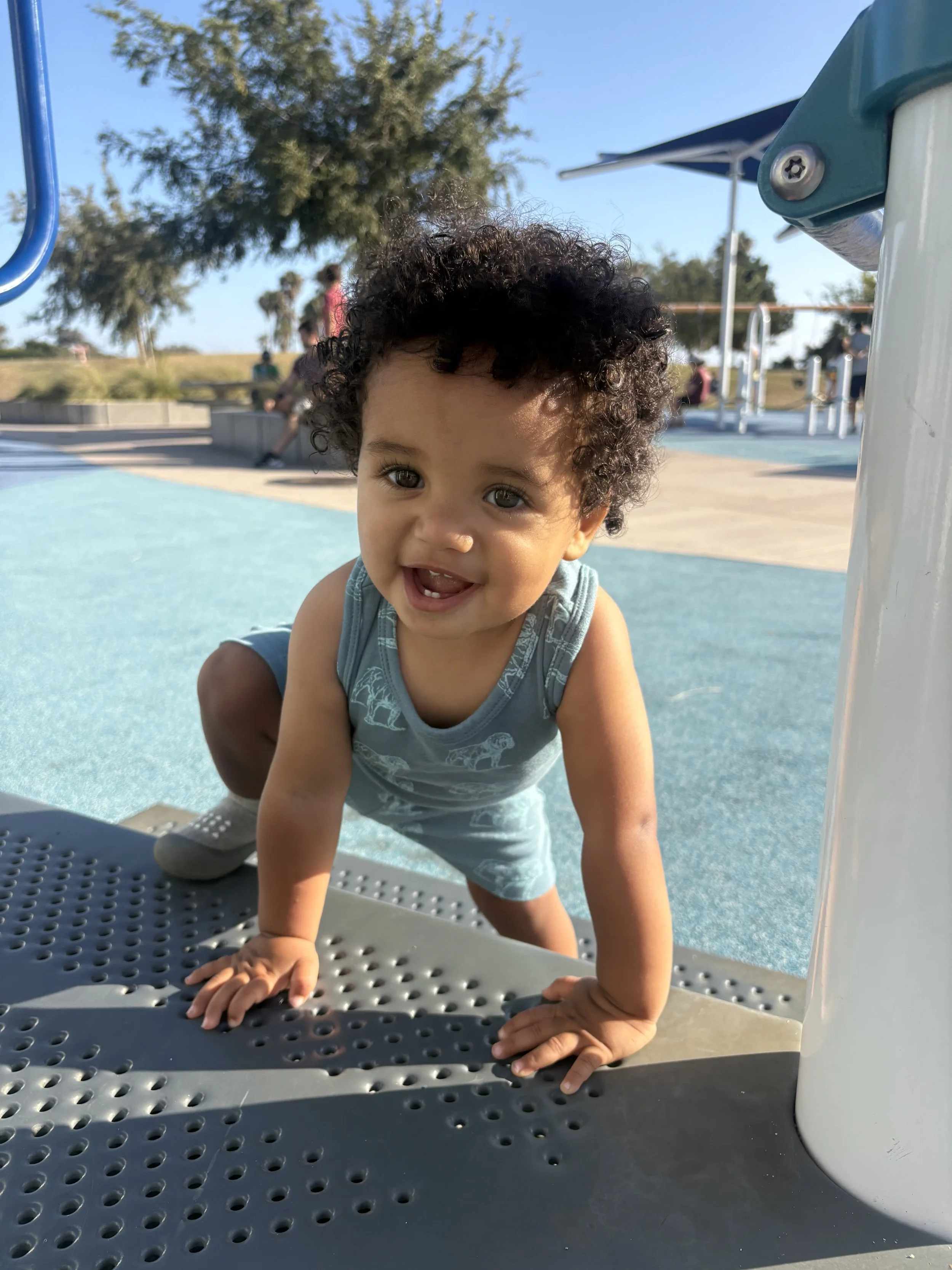 Young boy with curly hair smiling and crawling on a playground structure at a park during daytime.