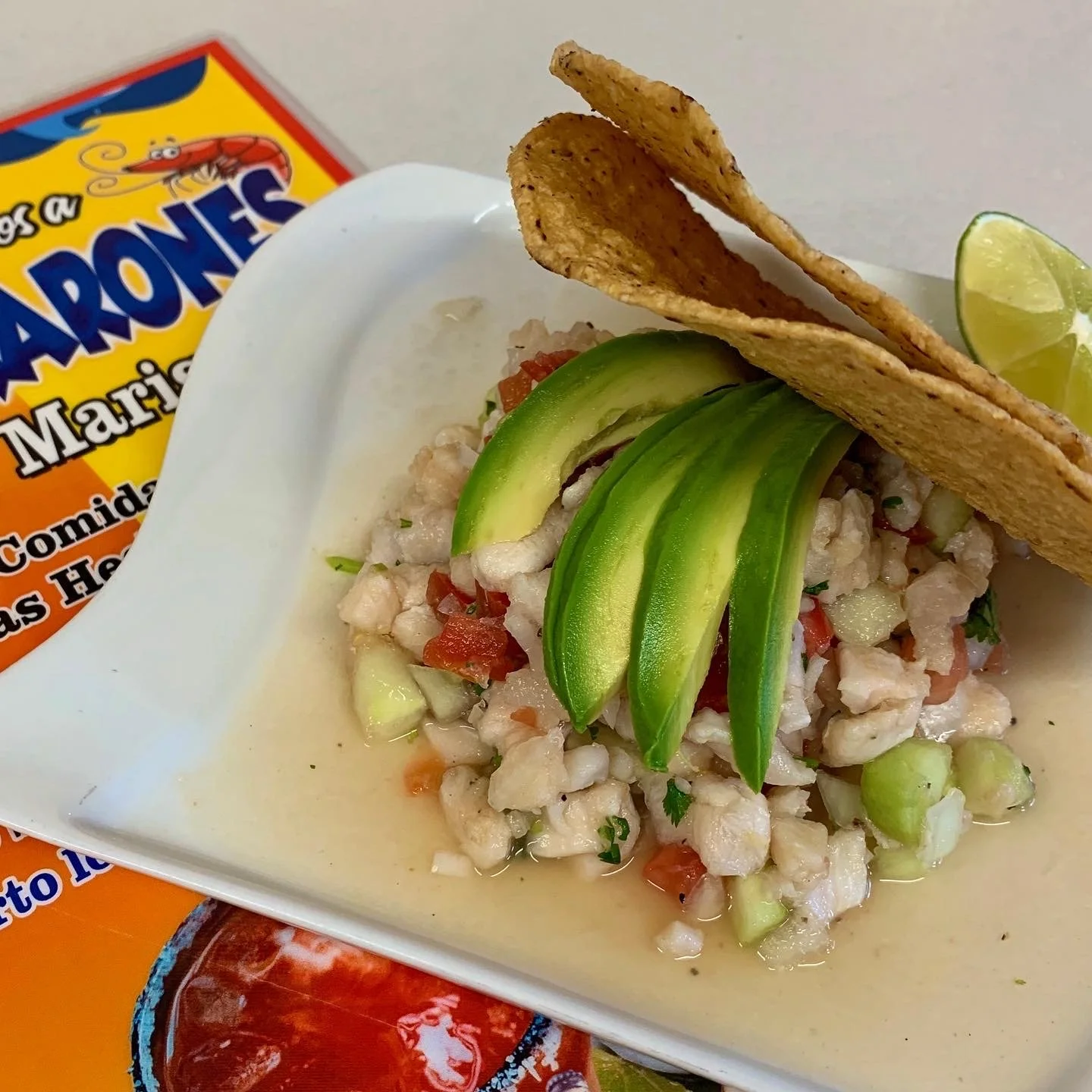 Close-up of a plate of ceviche with avocado slices, tortilla chips, and lime wedge, with a Paraisos Mariscos seafood brand advertisement in the background.