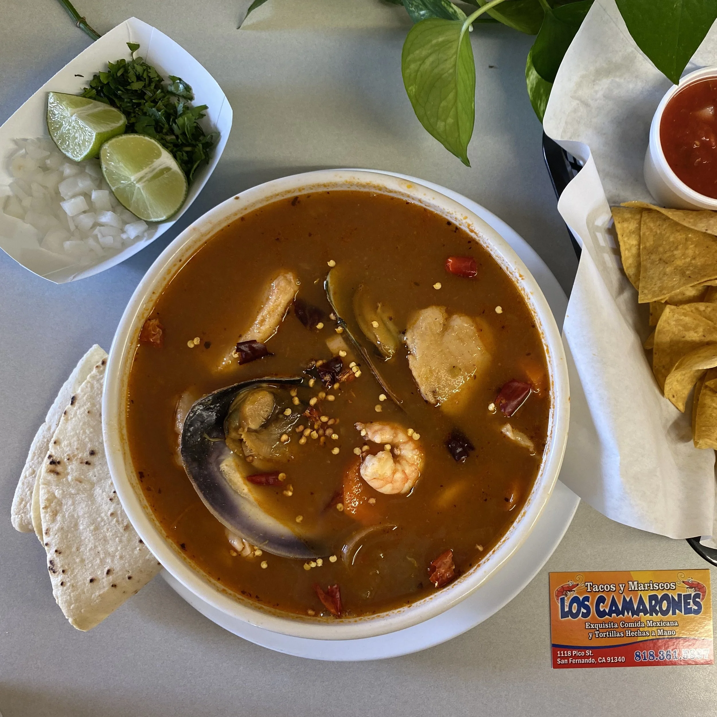 A bowl of Mexican seafood soup with shrimp, fish, vegetables, and broth, accompanied by a side of chopped onions, lime wedges, and chopped cilantro. A basket of bread and a small container of red salsa sit nearby on a gray table. A colorful restauran