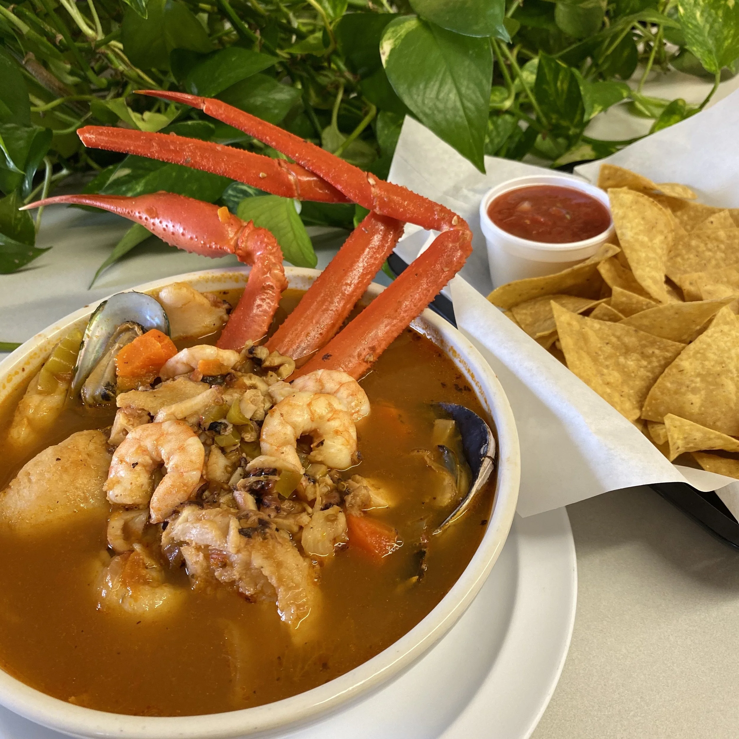 Bowl of seafood soup topped with crab legs, shrimp, and vegetables, served with tortilla chips and a side of salsa, with a backdrop of green leaves.