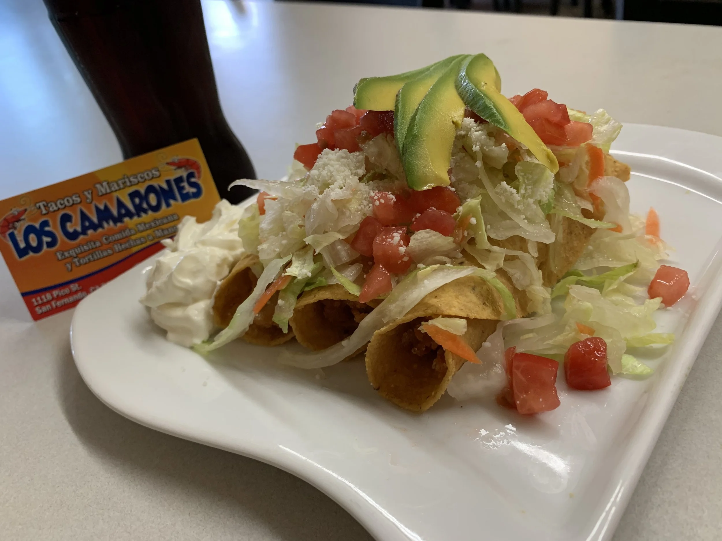A plate of Mexican tacos topped with chopped tomatoes, shredded lettuce, shredded cheese, and sliced avocado. In the background, a bottle of Los Camarones taco sauce is partially visible.
