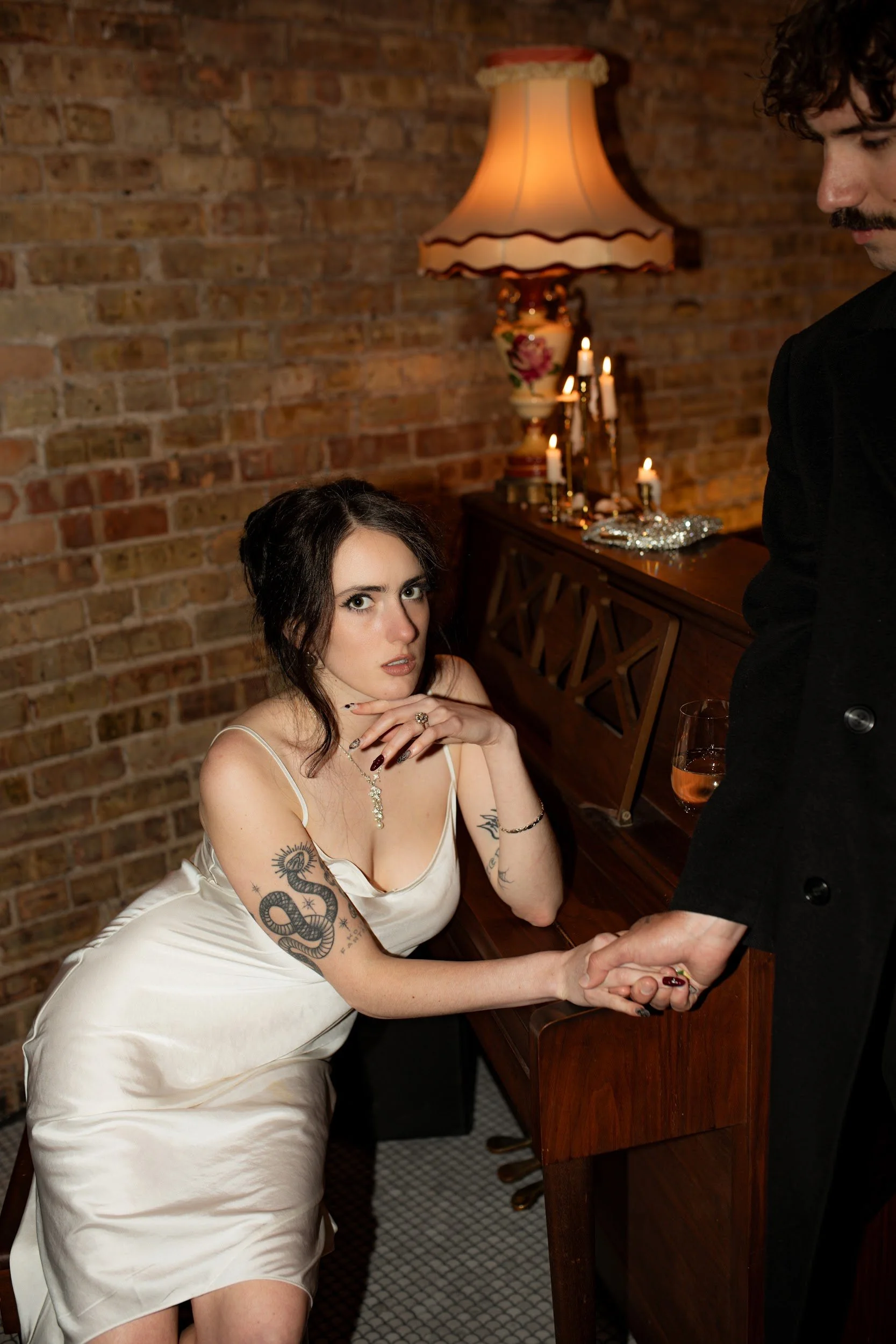 A woman in a white satin slip dress with tattoos on her left arm and chest, sitting at a bar, holding hands with a man who is standing. The background features a brick wall, a vintage lamp, and lit candles.