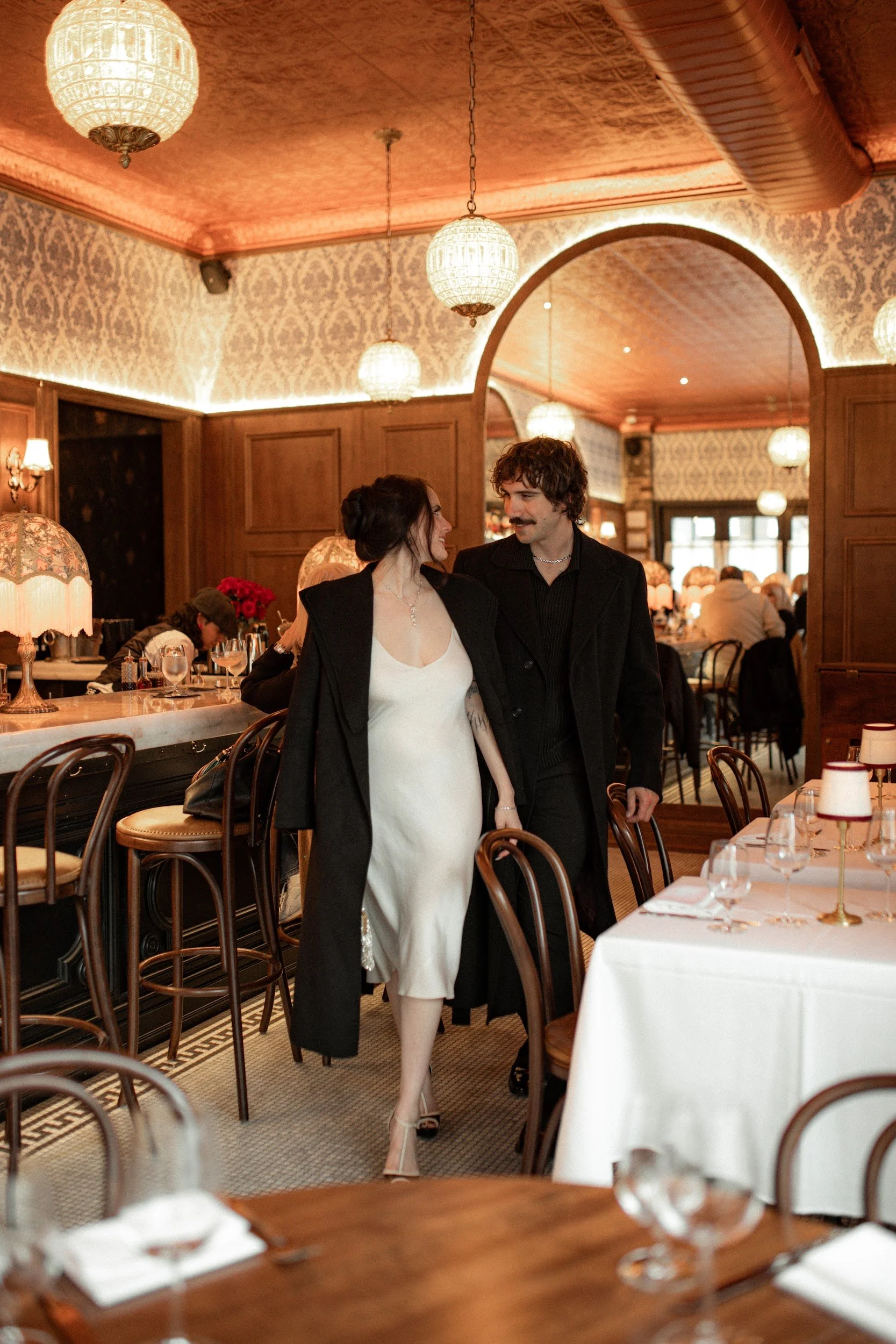 A man and woman are smiling at each other while walking in a restaurant with vintage decor, including wooden paneling, patterned wallpaper, and ornate lamps, during a formal or romantic dinner event.
