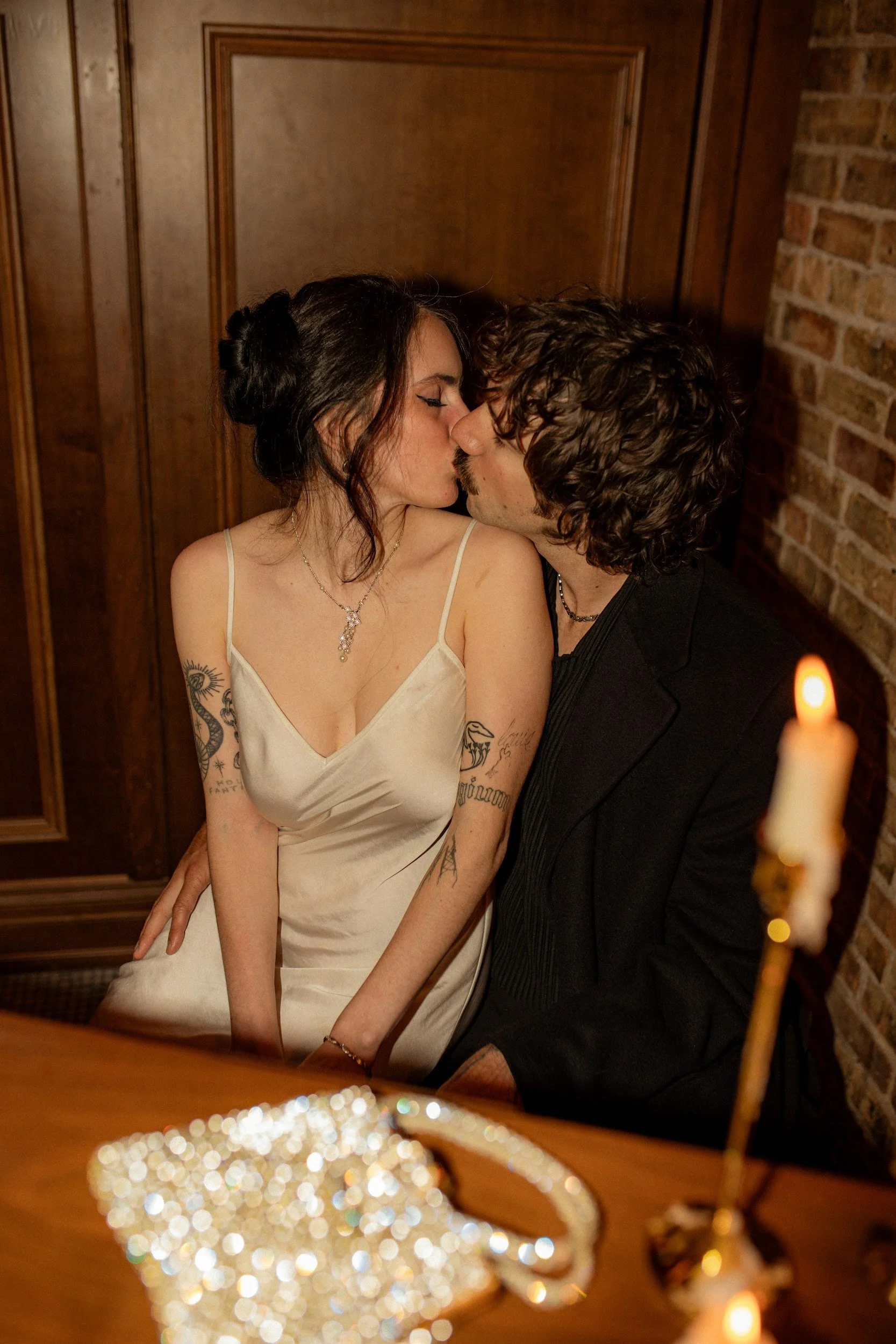 A couple kissing at a candlelit dinner, with a blurred centerpiece in the foreground and wooden paneled walls in the background.