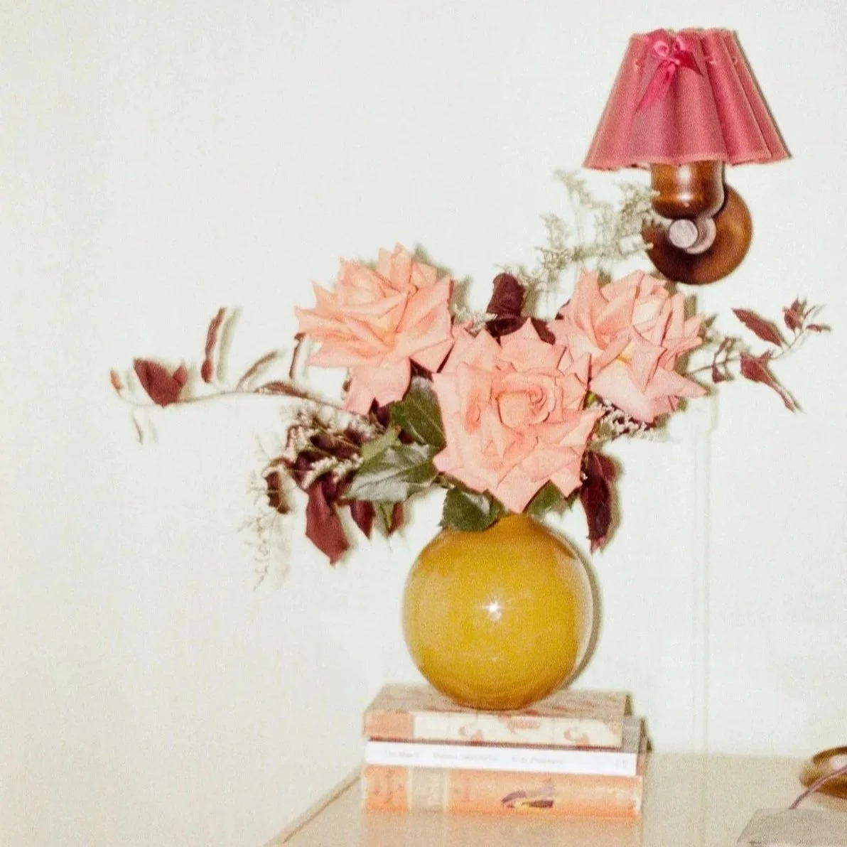A yellow vase with pink roses and foliage, placed on a stack of three books, with a pink lampshade attached to a wall-mounted lamp in the background.