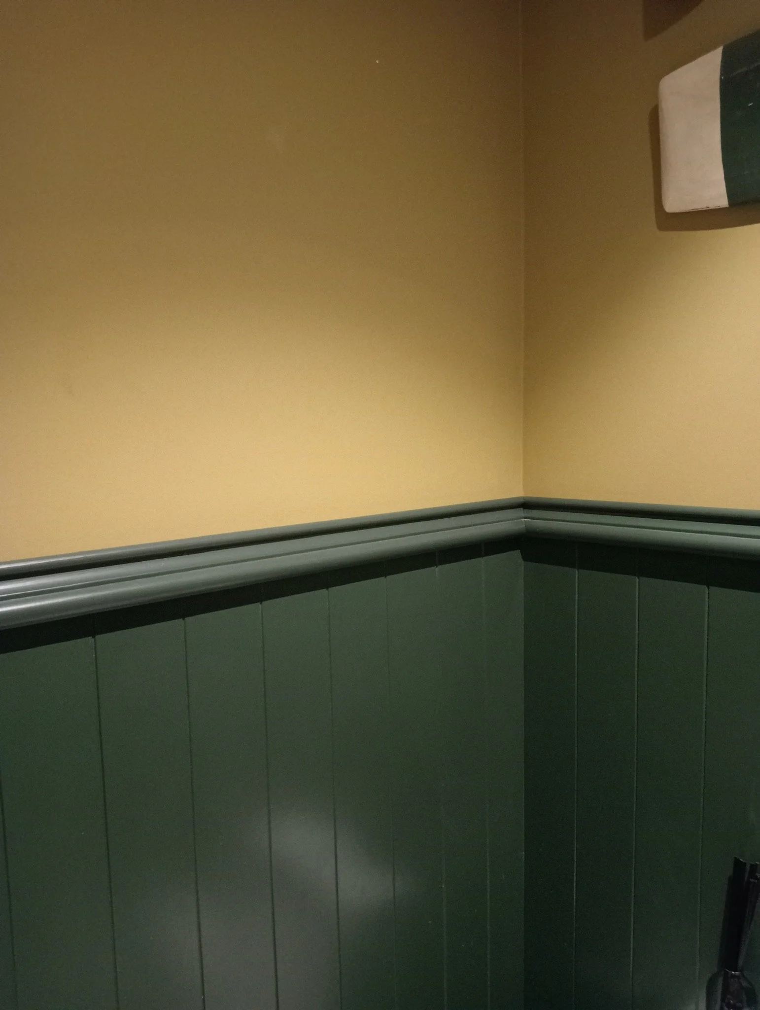 A corner of a room showing a beige wall, a green tiled lower wall, and a gray circular handrail. Part of a towel or cloth with green, white, and beige stripes is in the upper right corner.