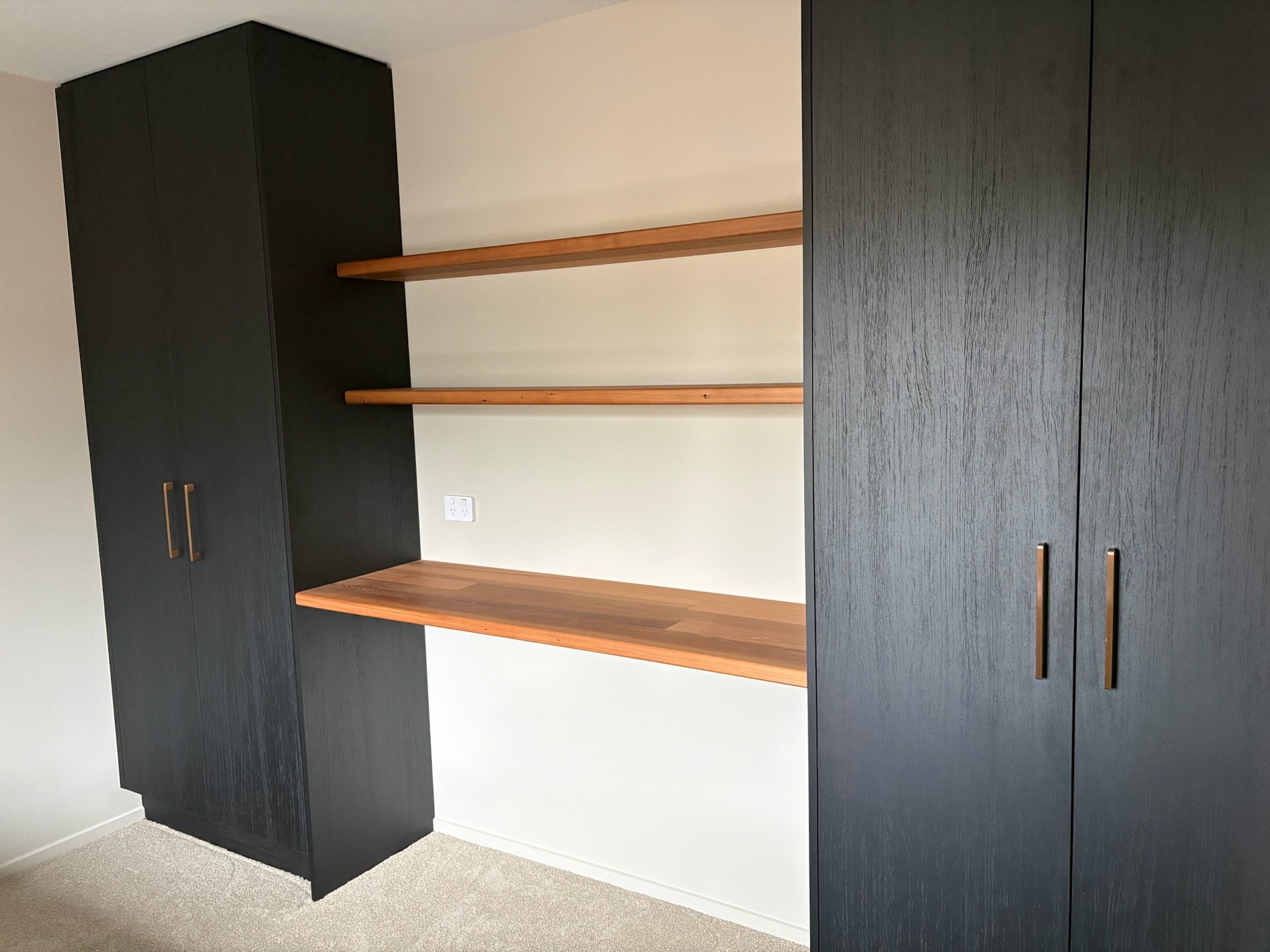 A modern black wardrobe with gold handles, open shelving with wooden shelves, and part of a matching black cabinet, set against a white wall with an electrical outlet.