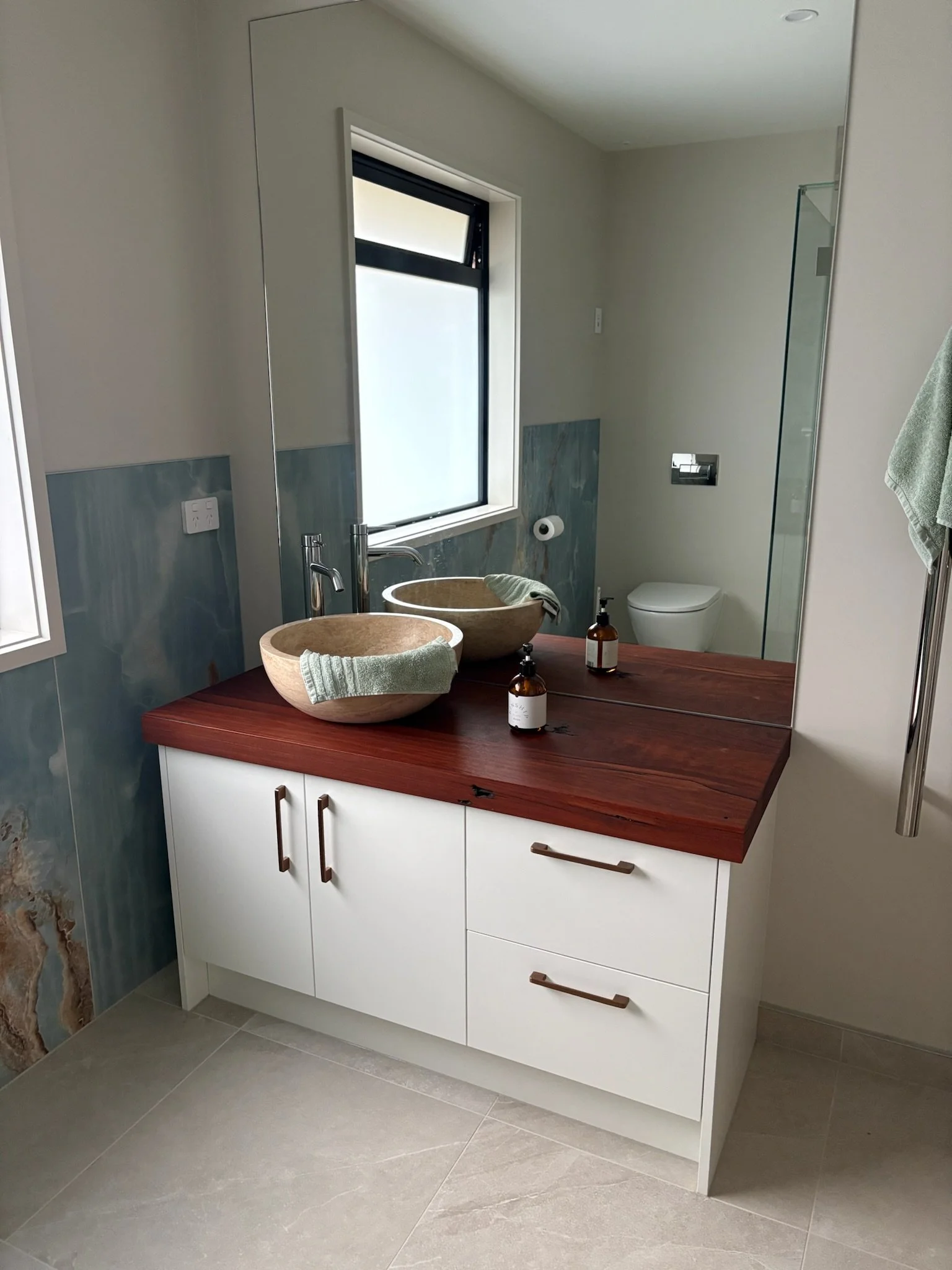 Modern bathroom vanity with white cabinetry, a dark wooden countertop, and two stone vessel sinks. A large mirror above the sinks reflects a window and part of a shower enclosure. Toiletries and towels are on the countertop.