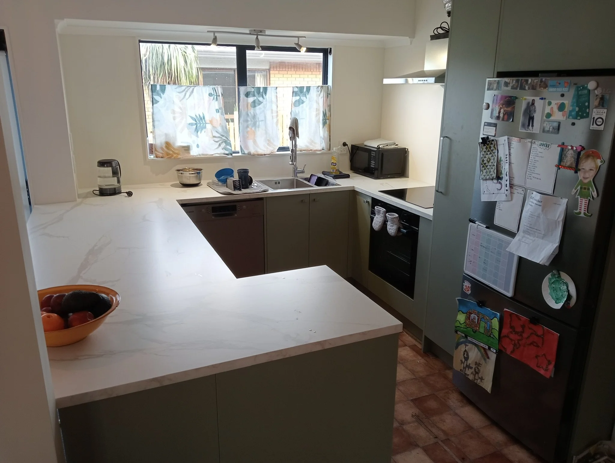 A kitchen with a marble countertop, a window with floral curtains, and various appliances on the counter including a microwave, small black appliance, and a thermos. There are children's drawings and photos on the fridge, and a bowl of fruit on the countertop.