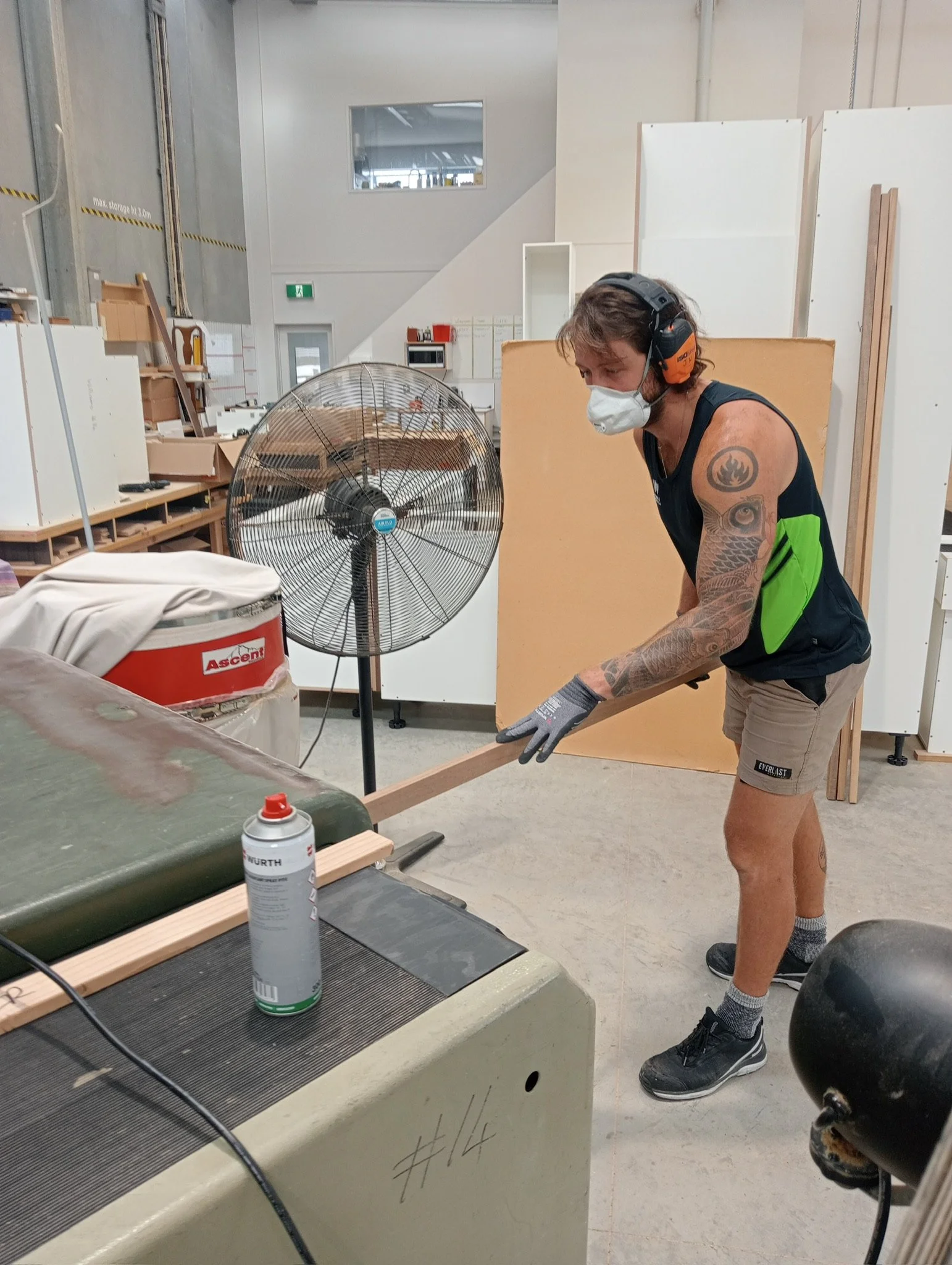 A man in a sleeveless shirt, shorts, and gloves working with a piece of wood in an industrial or workshop setting. He is wearing a white mask and noise-canceling headphones. There is a fan and a container of spray on the work surface, with shelves and equipment in the background.