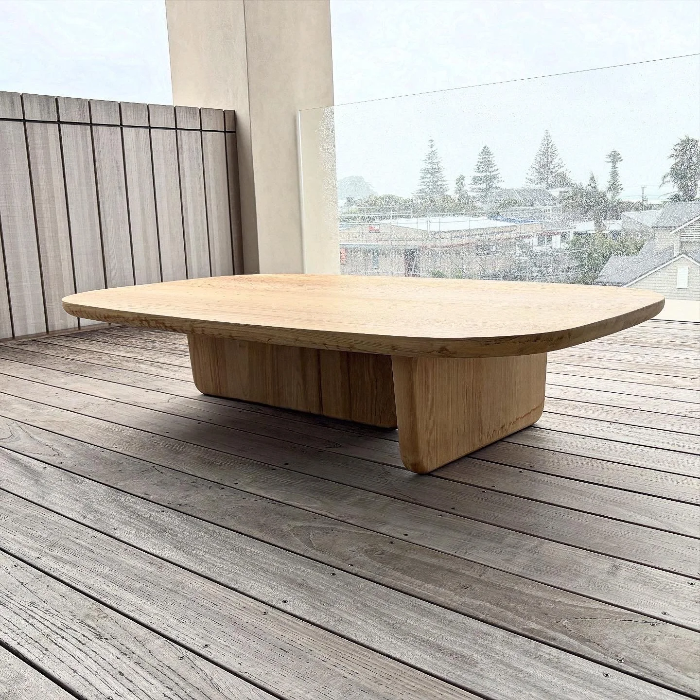 A low, modern wooden table on a wooden deck with a backyard and cloudy sky in the background.