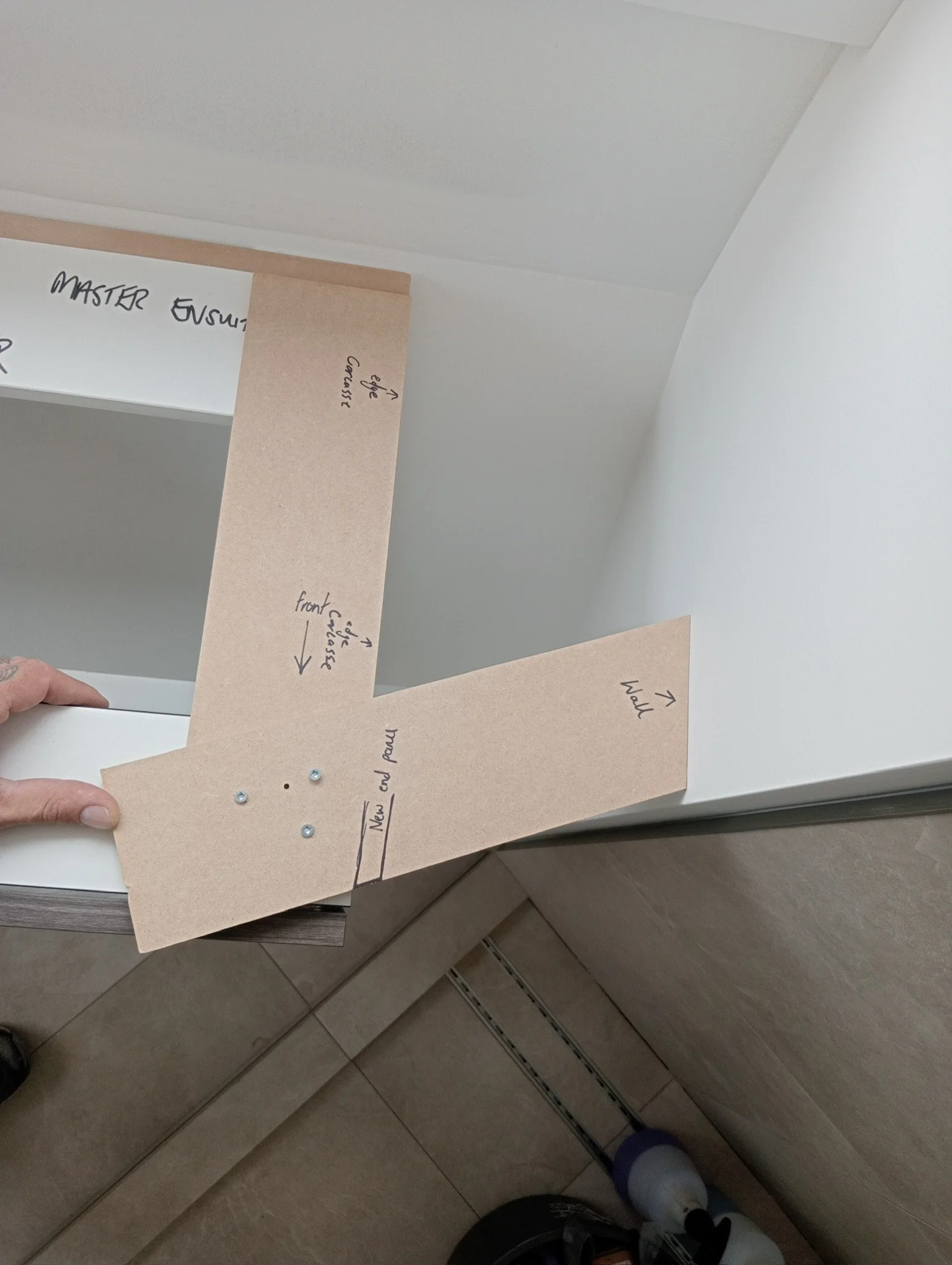 A person is holding a piece of tan construction paper with handwritten notes and arrows, which appears to be part of a project or craft. The paper has labels like 'MASTER', 'Front', and 'WALL'. Behind the paper, there is a mirror reflecting the ceiling and part of the room.