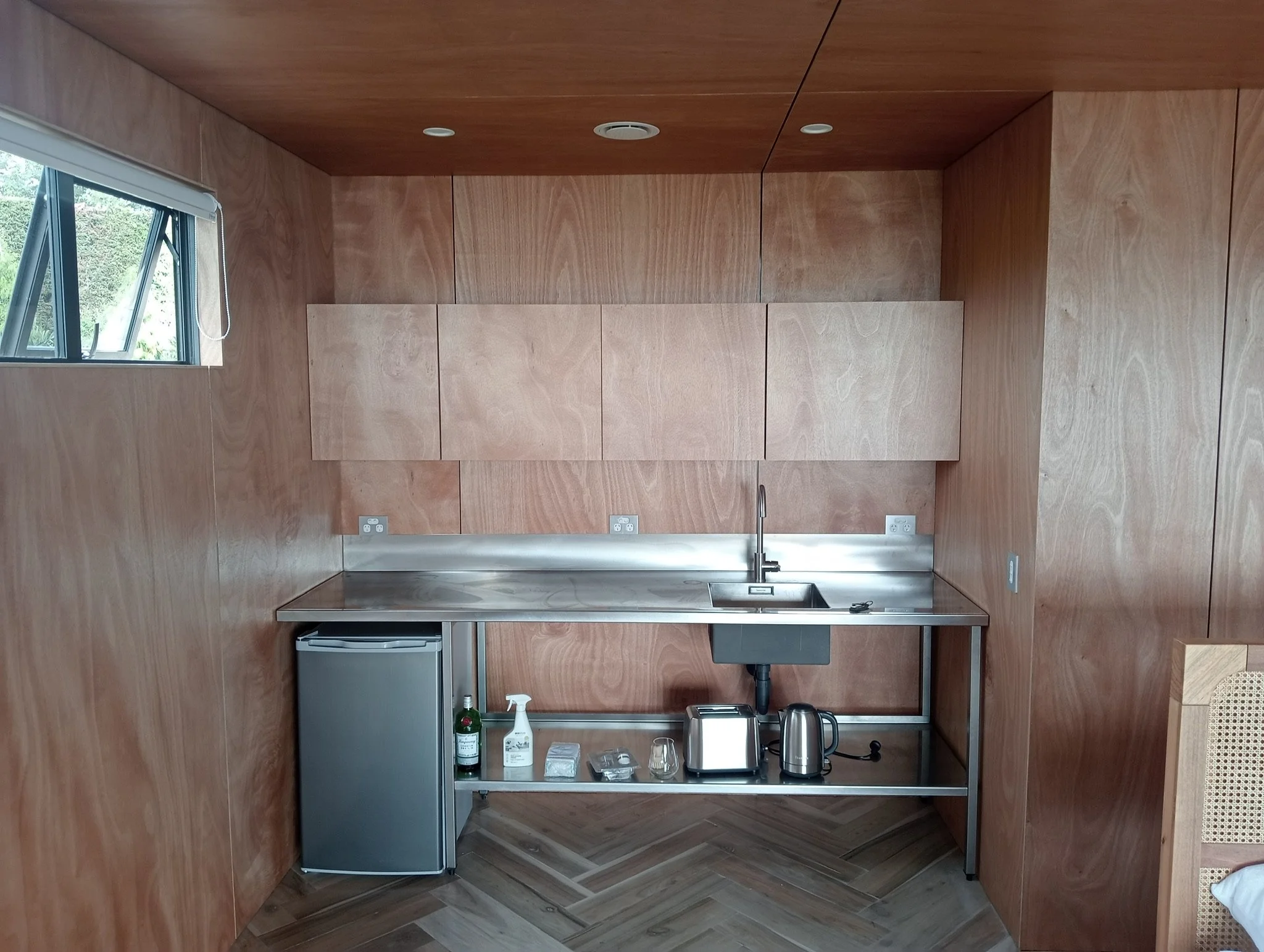 A kitchen with wood-paneled walls and ceiling, a small window, a stainless steel sink, a mini fridge, a kettle, two electric kettles, a soap dispenser, and various small items on a lower shelf.