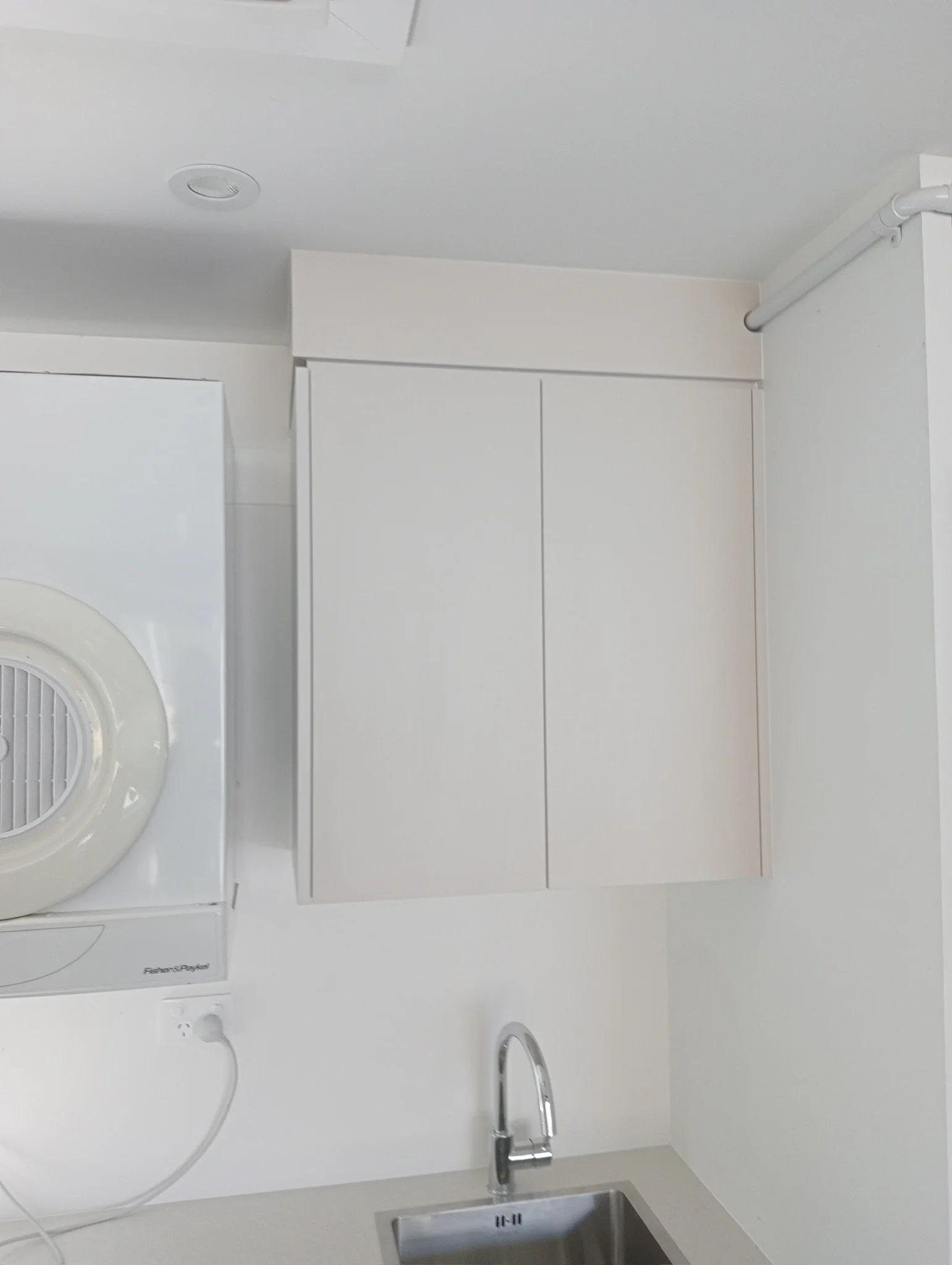 A white laundry area with a Fisher & Paykel washing machine, a white tiled backsplash, a sink with a stainless steel faucet, and a white cabinet above the sink.