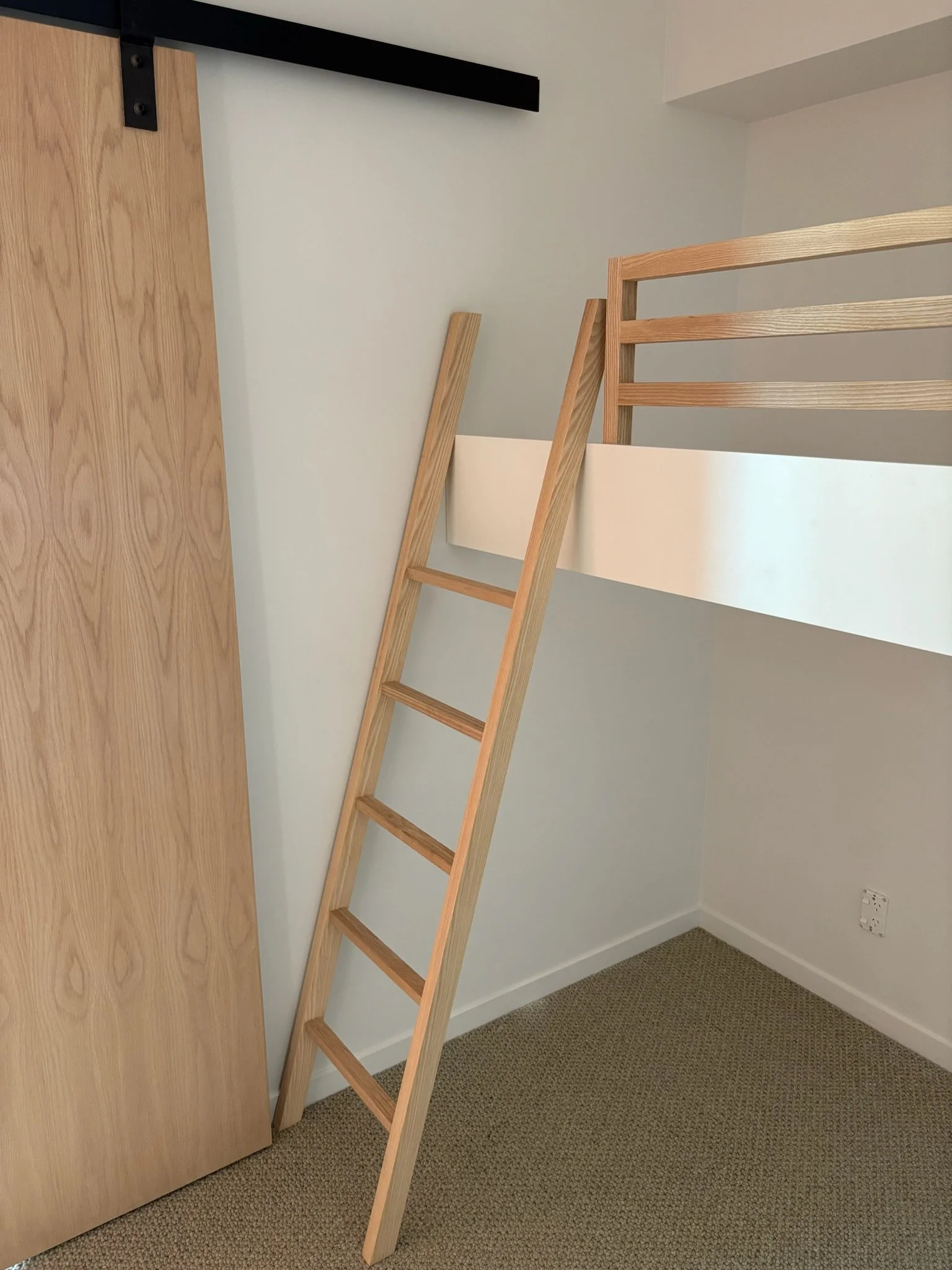 A wooden ladder leaning against a white wall in a room with beige carpet and a wooden door.