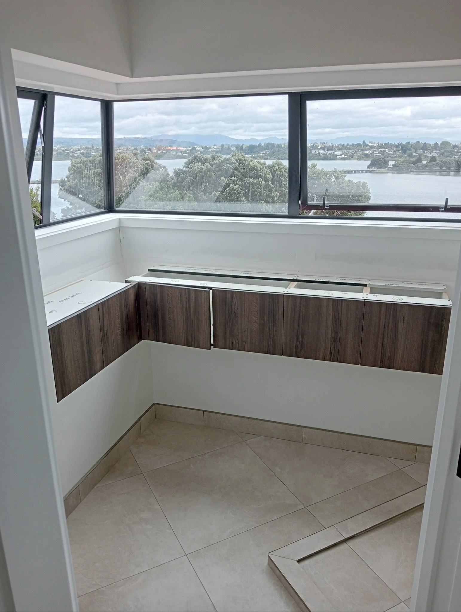 Inside view of a small corner space with large windows overlooking a lake, trees, and a distant landscape, and kitchen cabinetry under construction with countertops yet to be installed.