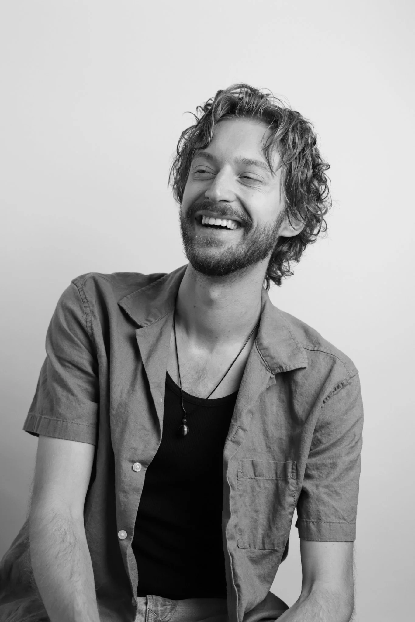 Black and white photo of a man with curly hair and a beard, smiling and looking slightly to his right, wearing a short-sleeved shirt and a necklace, against a plain light background.