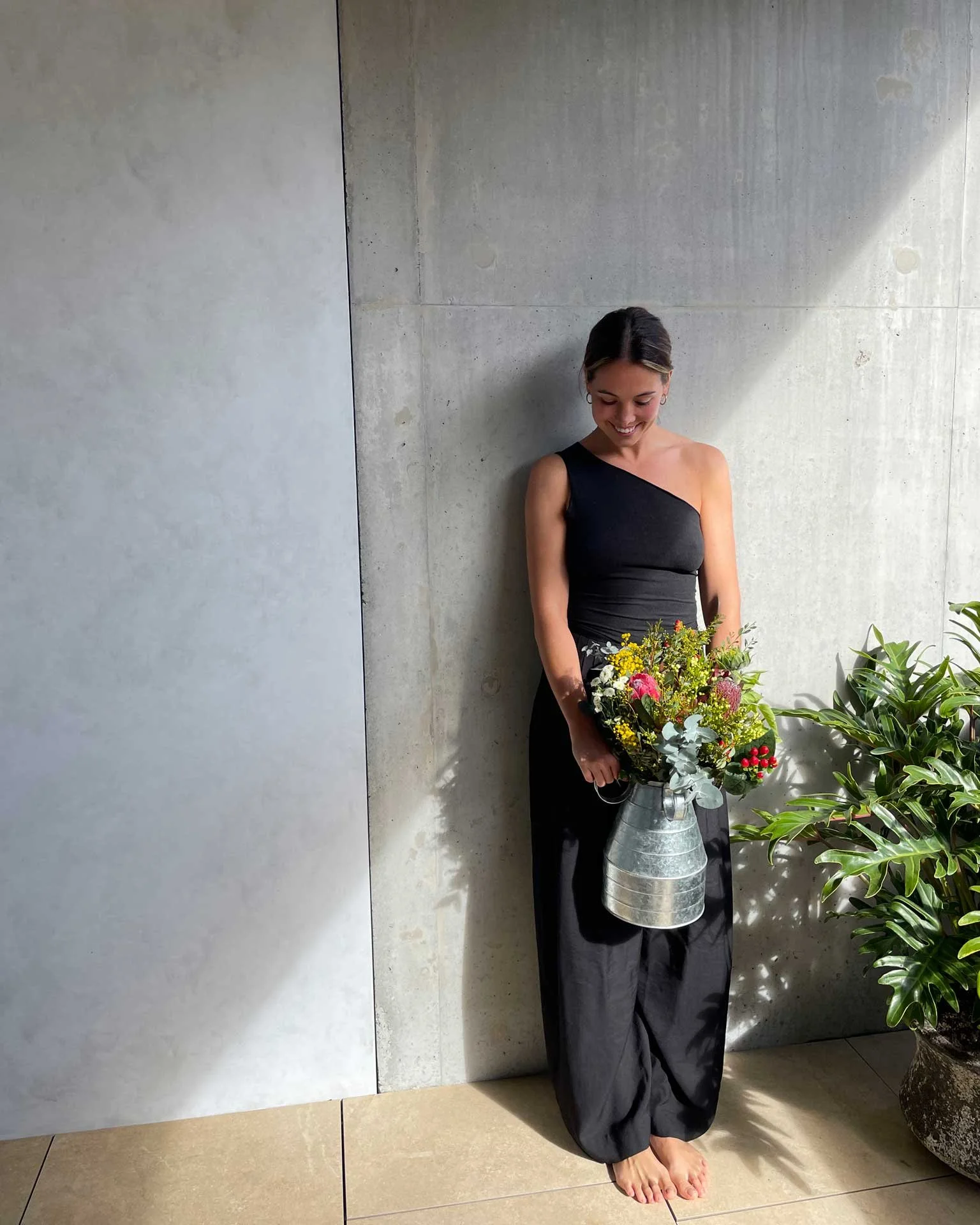 A woman in a long black dress with one-shoulder design, standing barefoot next to a concrete wall, holding a metal watering can filled with colorful flowers, with potted plants nearby.