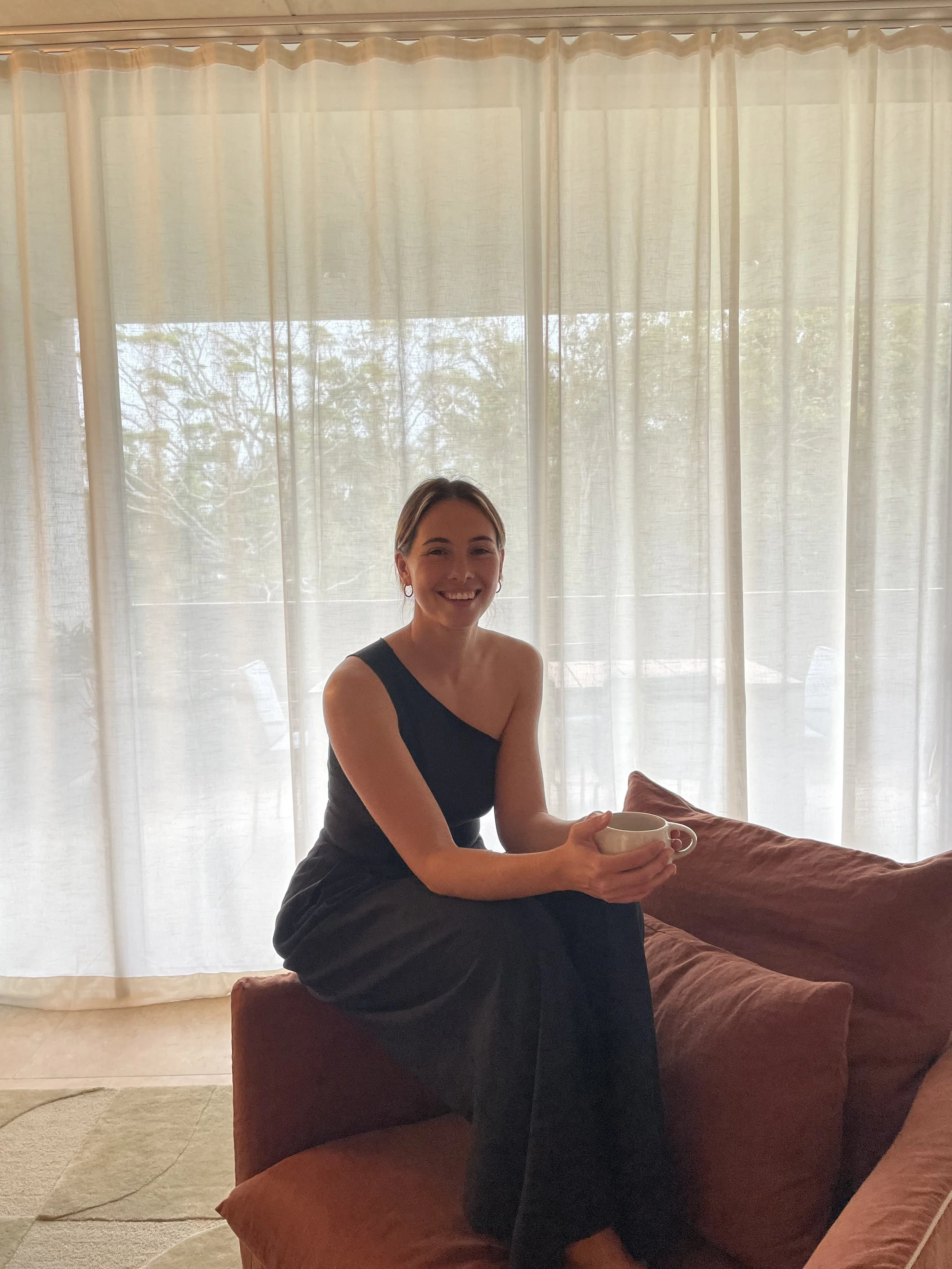 A woman sitting on a brown couch, holding a mug, smiling in front of a large window with white curtains, with trees visible outside.