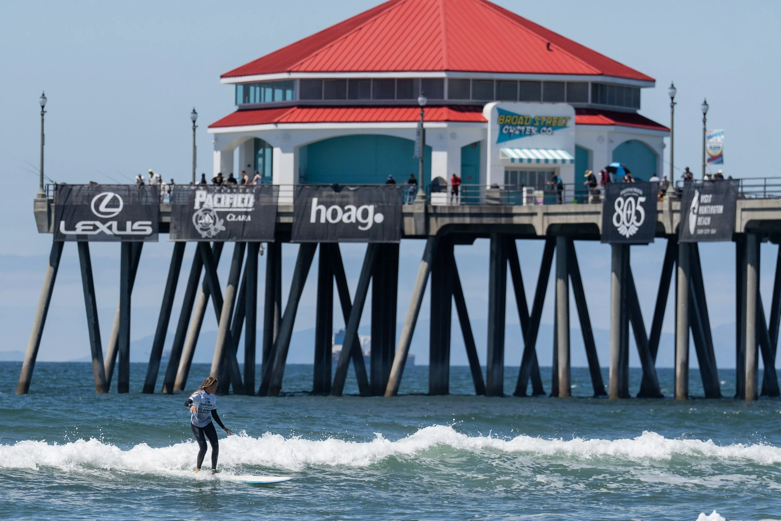 A person surfing in the ocean near a pier with a red-roofed building and banners for Lexus, Pacifica, Hoag, and 805 in the background.