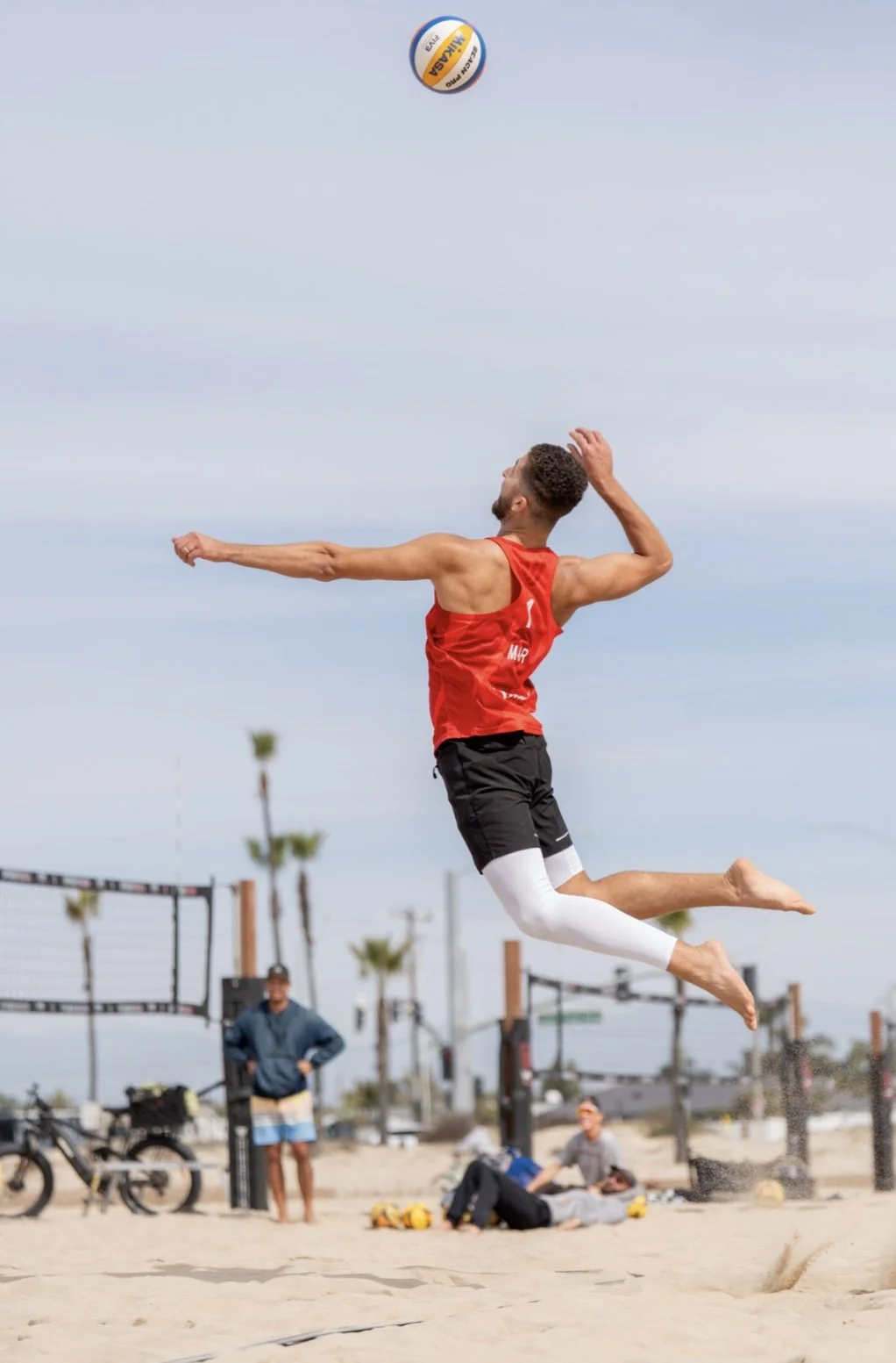A man in athletic clothes jumping to hit a volleyball on a beach with a volleyball net and people in the background.