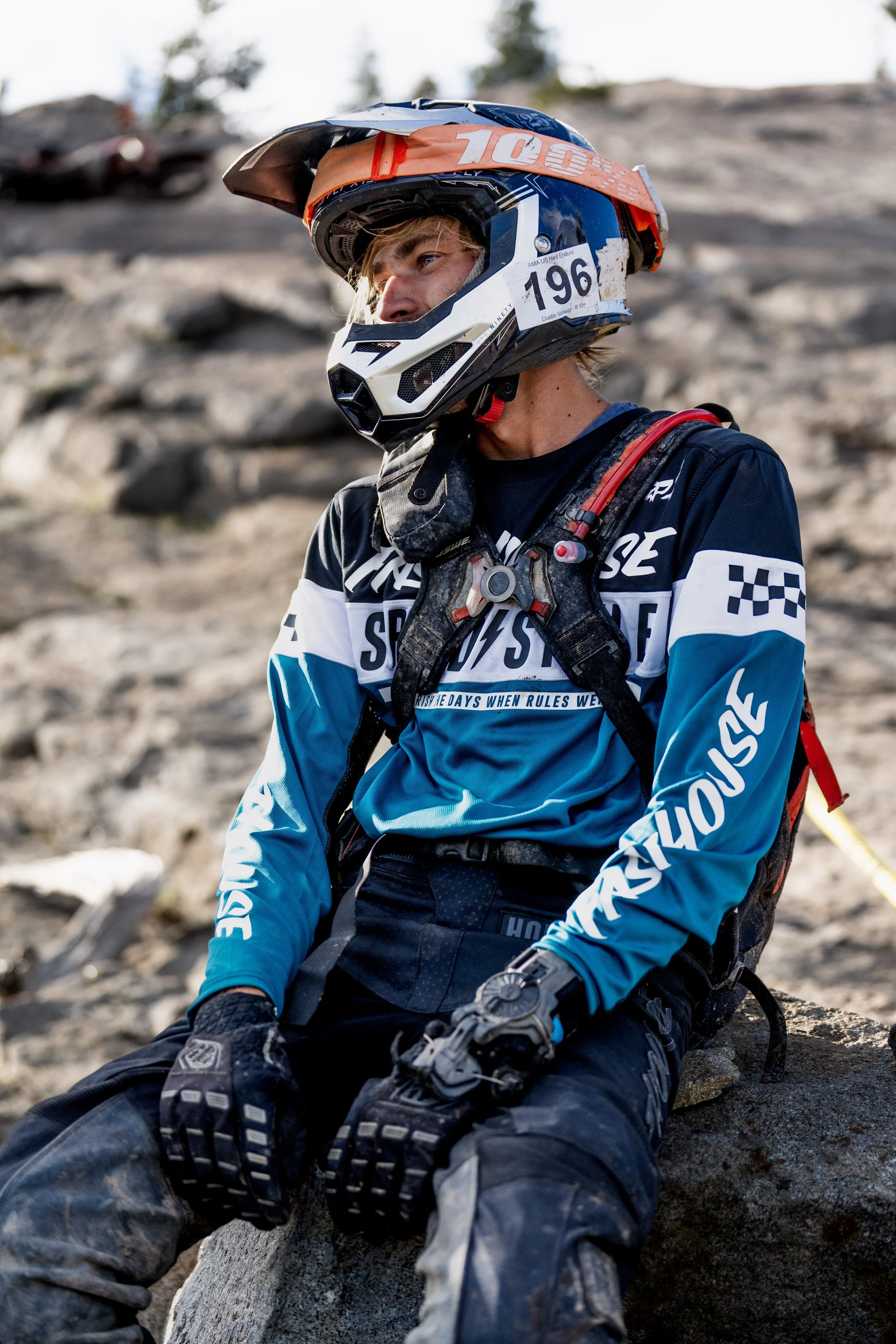 A person wearing a motocross helmet, goggles, and gear sitting on a rocky terrain with a blurry background of rocks and trees.