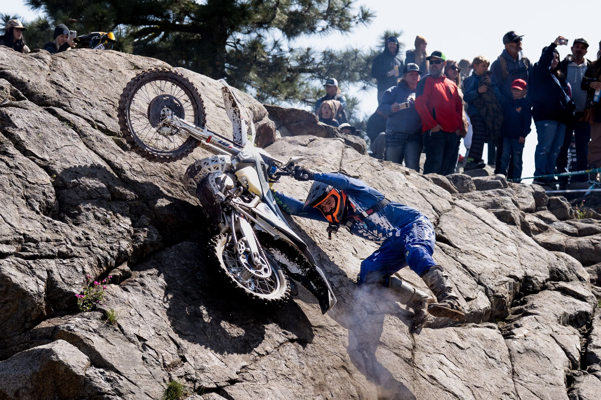 Someone falls while mountain biking on a rocky trail, while a group of onlookers watches and records the incident.