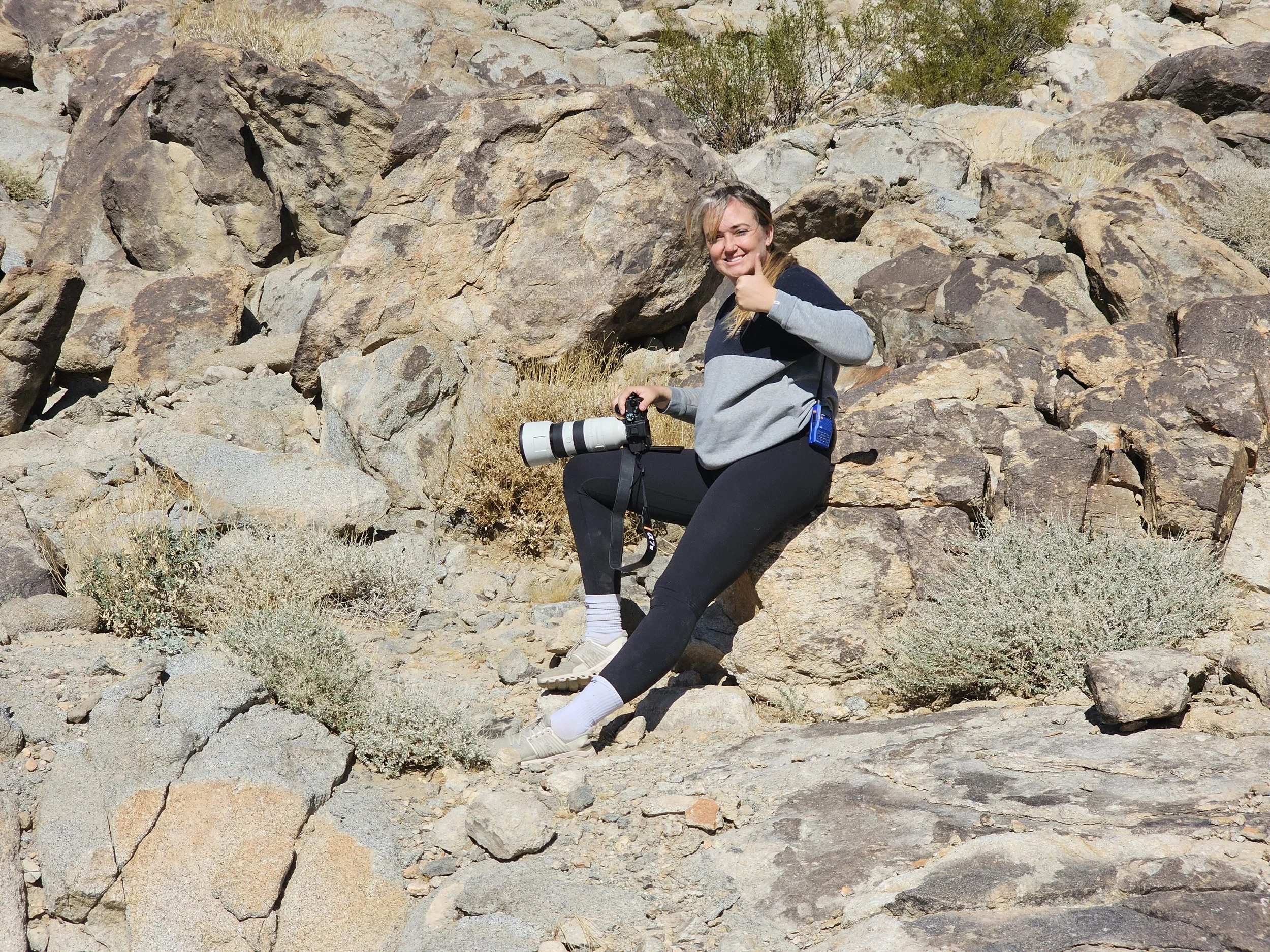 A woman smiling and giving a thumbs-up, sitting among rocks in a desert landscape with sparse vegetation, holding a camera with a large telephoto lens.
