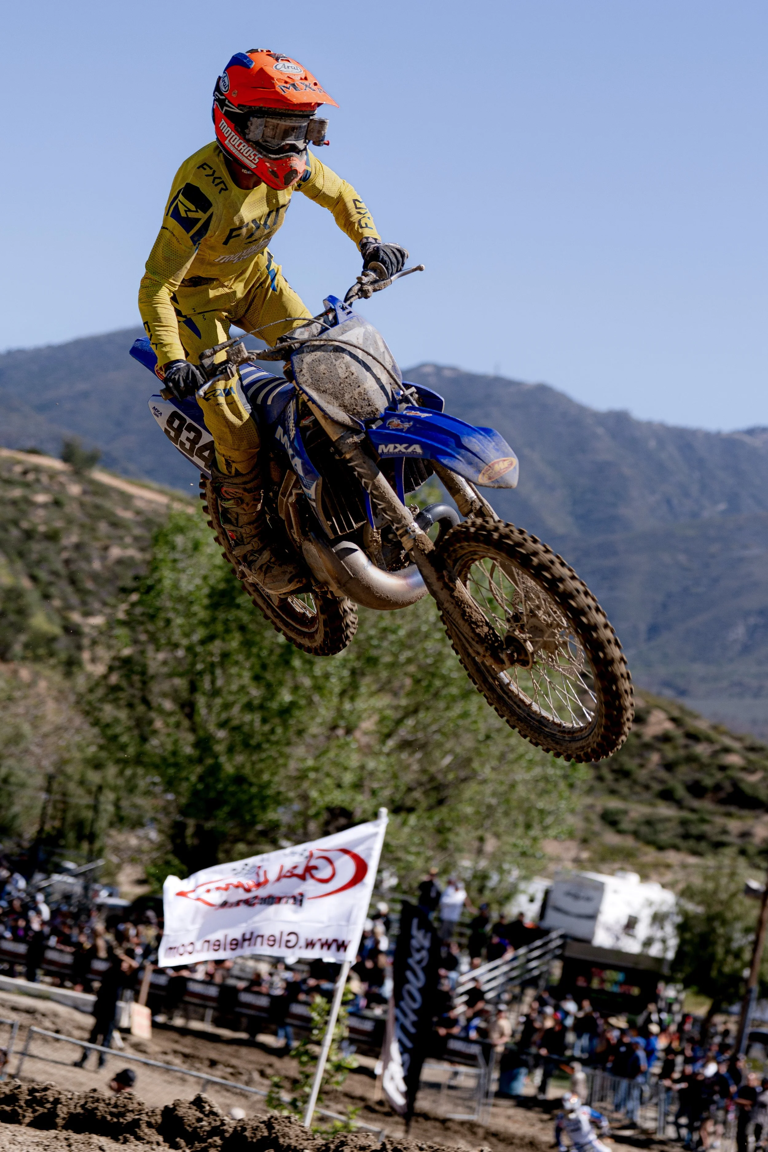 A motocross rider wearing a yellow suit, red helmet, and goggles jumping mid-air on a blue dirt bike over a dirt track with a crowd and banners in the background against a mountainous landscape and clear blue sky.