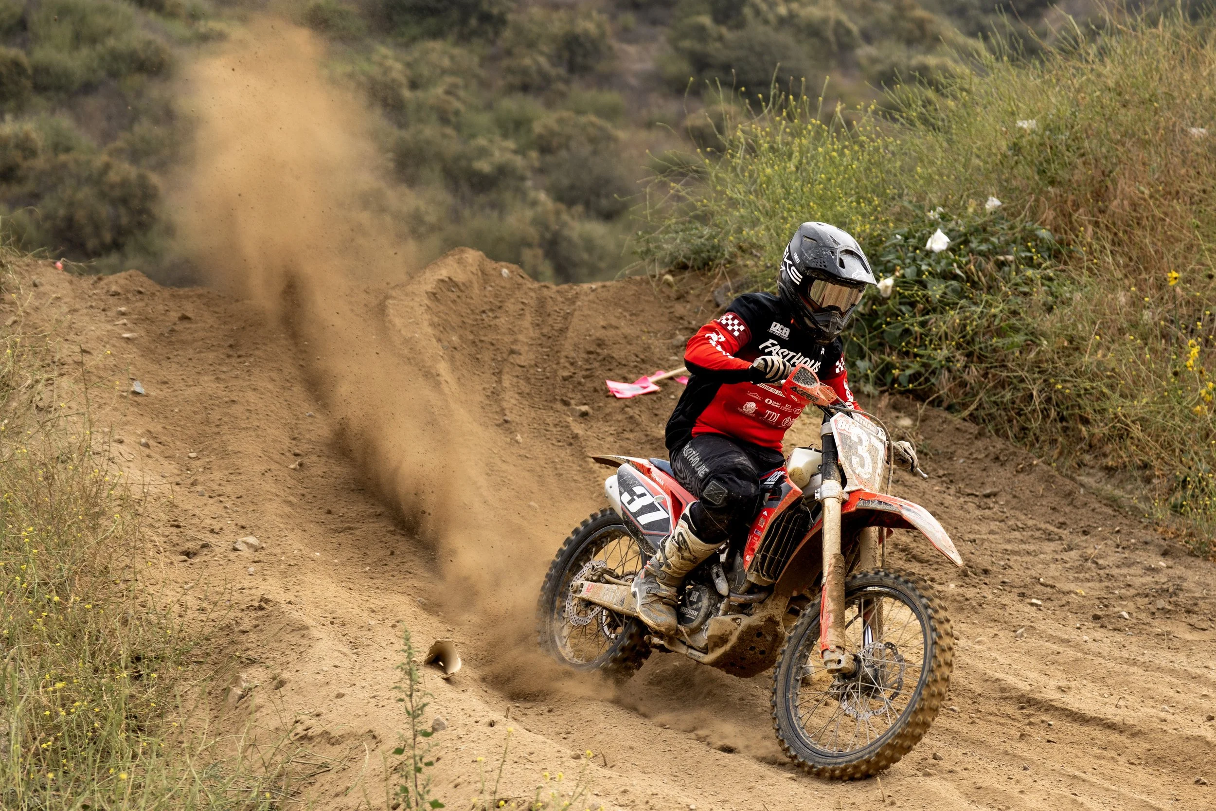 A motocross rider in red and black gear and a helmet with goggles riding a dirt bike on a dirt trail, kicking up dust and dirt with a hillside and shrubs in the background.