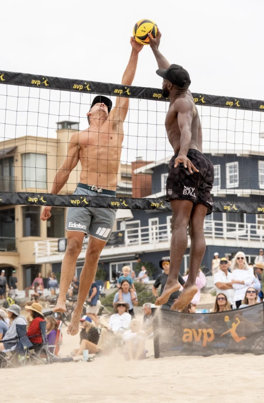 Two male beach volleyball players jumping at the net, one hitting the ball over the net and the other attempting to block, with an audience watching in the background.