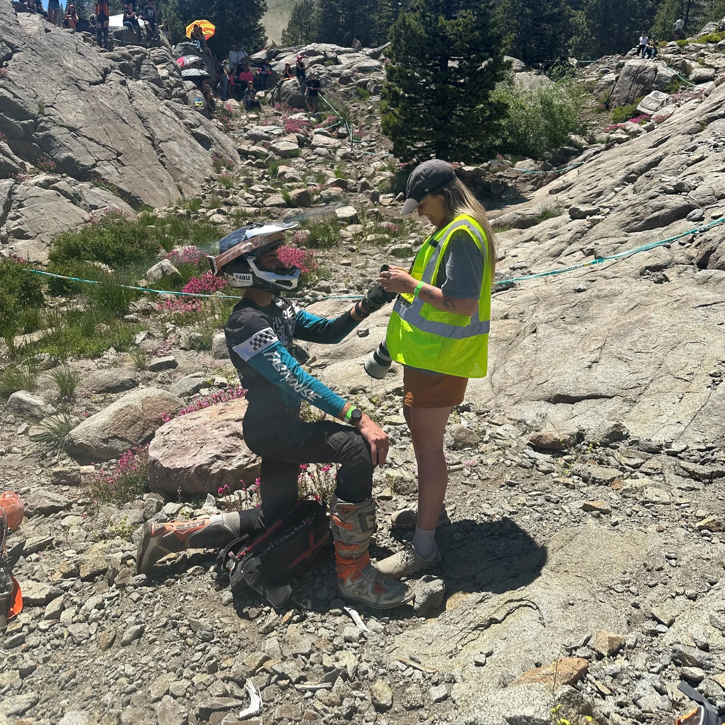 A person in mountain biking gear kneels on rocky terrain while talking to a woman in a yellow safety vest and brown shorts, in a mountainous area with other people perched on rocks and trees in the background.