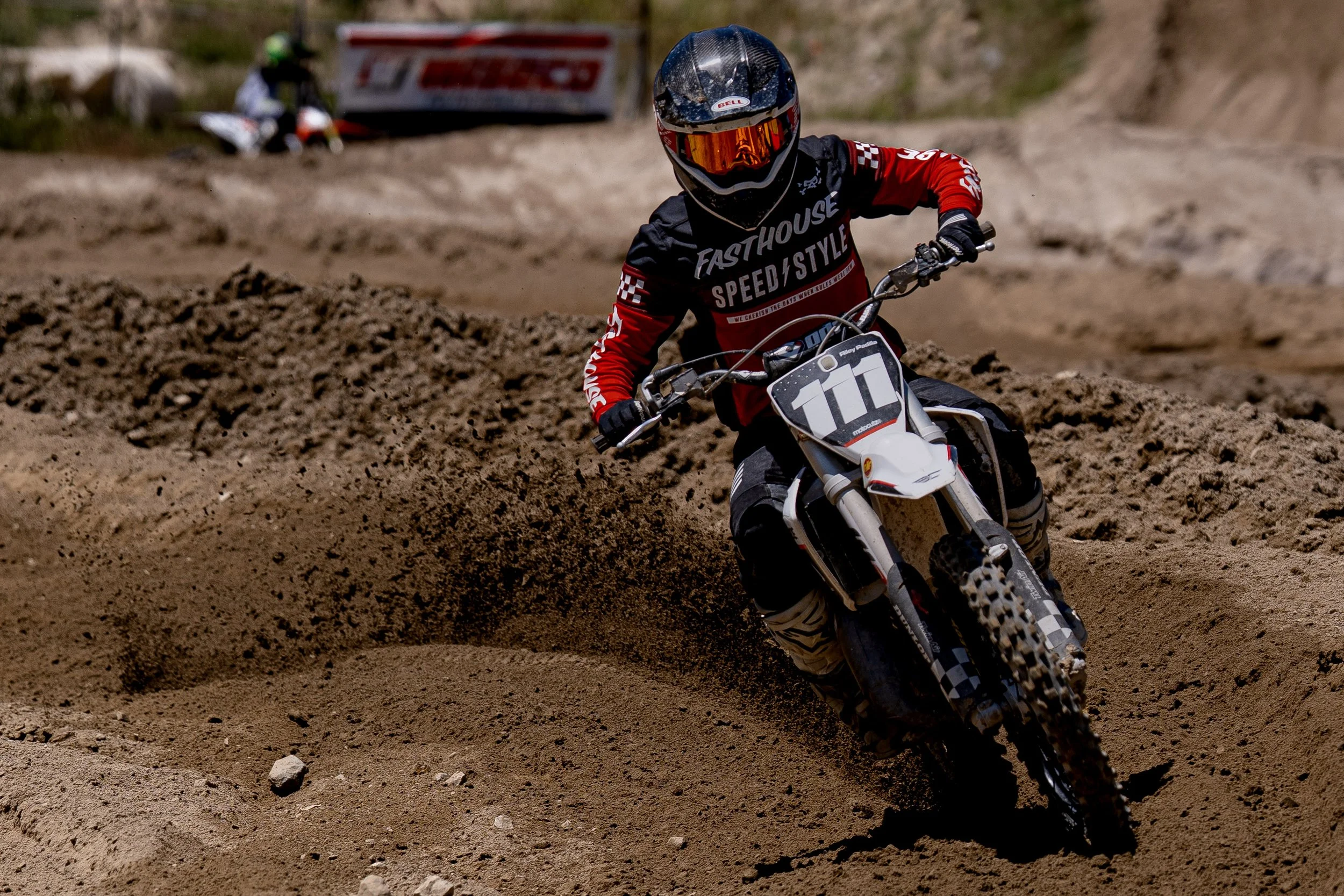 A motocross rider wearing a black helmet with orange visor, black and red gear with 'FastHouse' and 'SpeedStyle' logos, racing on a dirt track. Another rider on a motorcycle is visible in the background.
