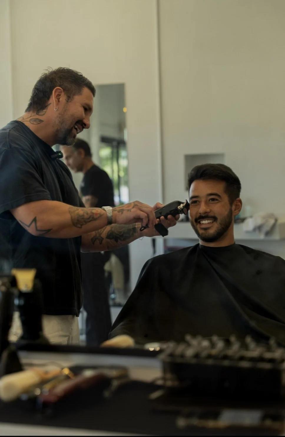 A man with a beard and short dark hair is sitting in a barber shop chair, smiling, while a barber with tattoos and earrings uses clippers to trim his hair. The barber has short dark hair, a beard, tattoos on his arms and neck, and wears a black shirt