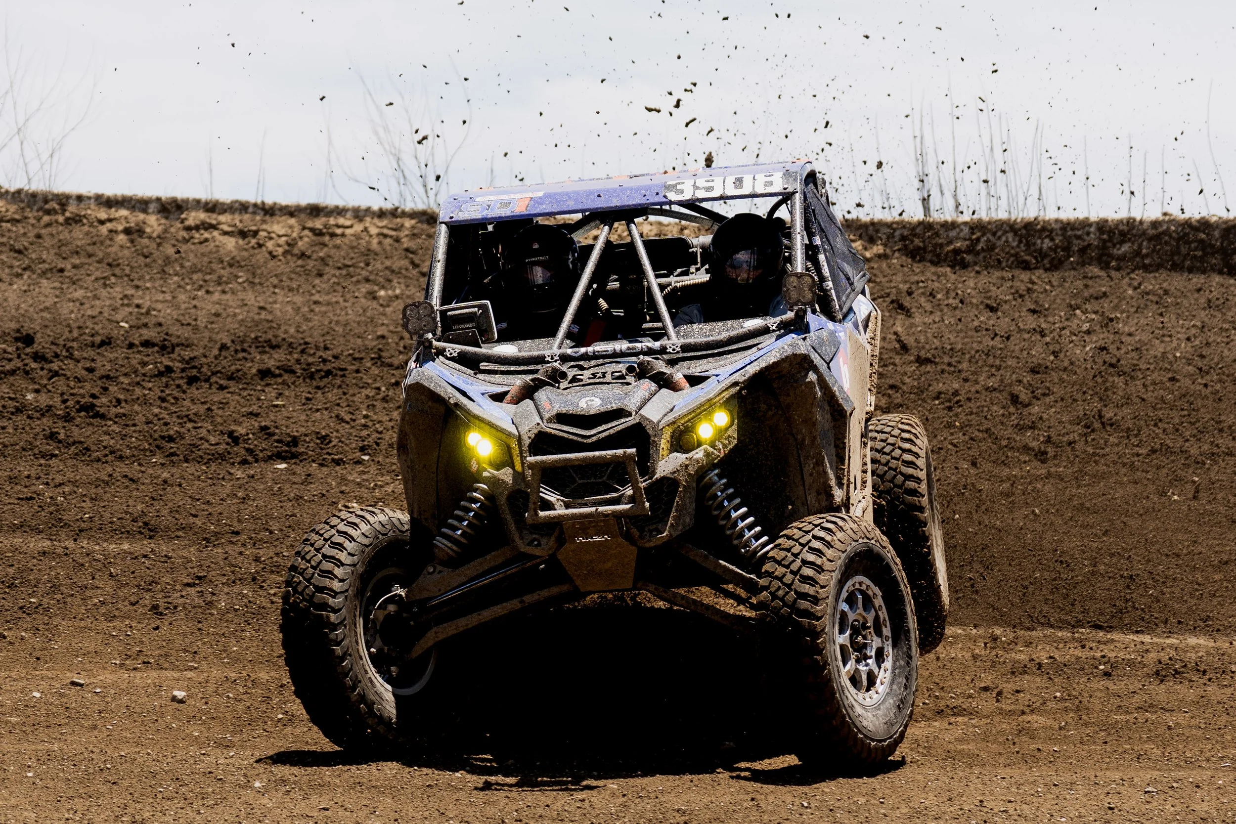 A blue off-road vehicle kicking up dirt while driving on a dirt track with a cloudy sky in the background.