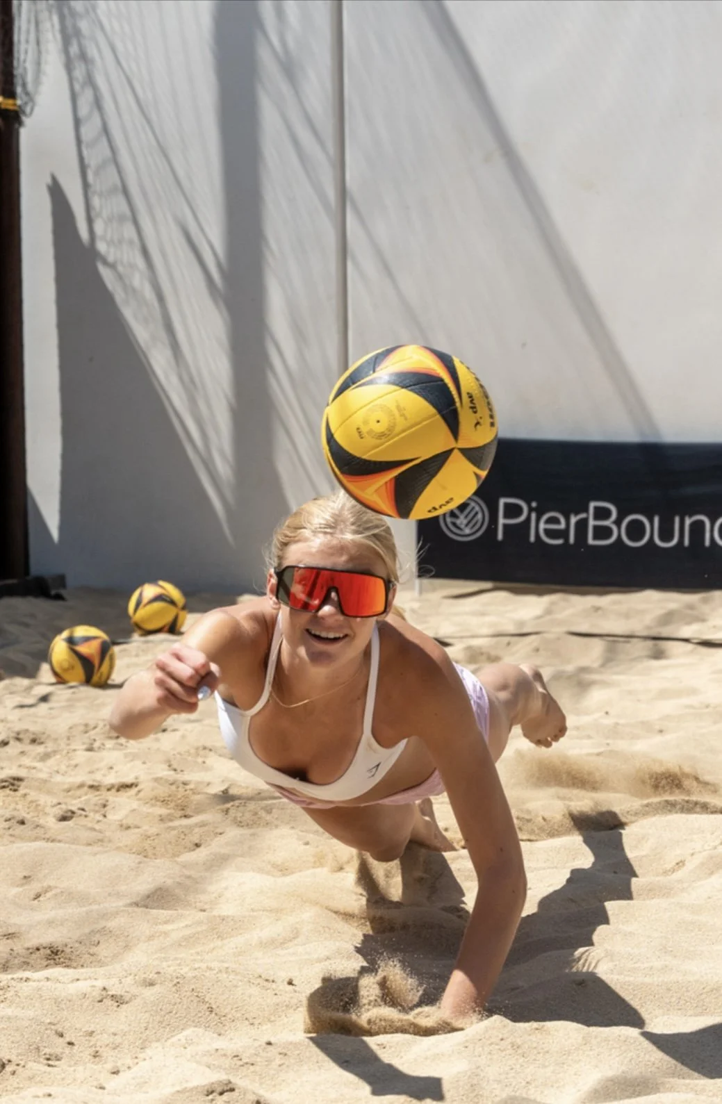 A woman in a white sports bra and shorts, wearing red sports sunglasses, is playing beach volleyball on sand. She is diving forward, reaching out with her right hand, and smiling. Several volleyballs are on the sand behind her, and a large volleyball