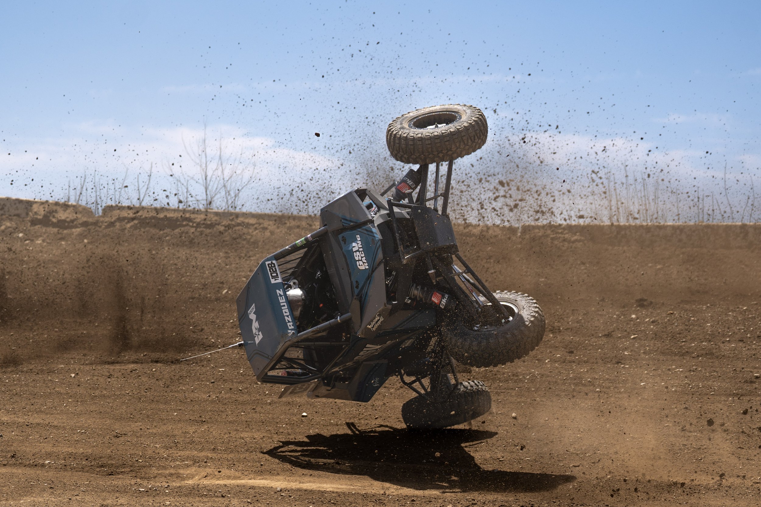 An off-road vehicle flips over on a dirt track during a dirt bike race, with dirt and mud flying in the air.