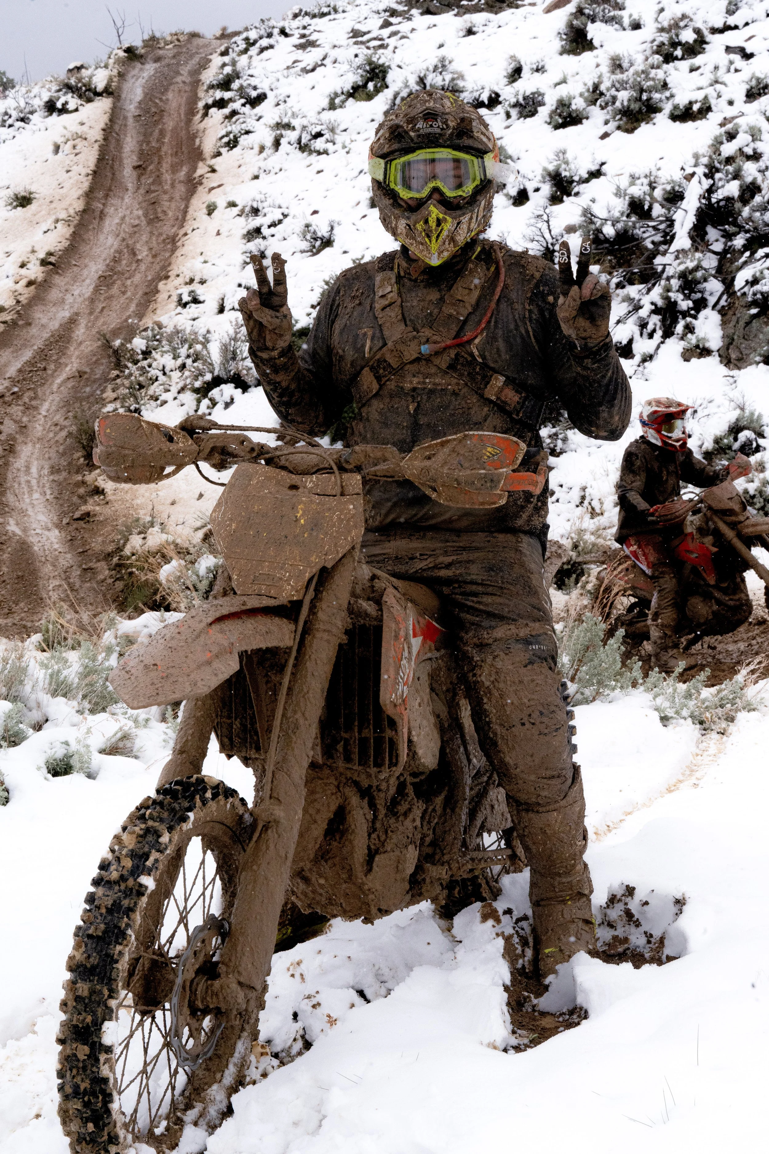 Off-road dirt bikes covered in mud riding on a snowy trail in a hilly, snow-covered landscape.