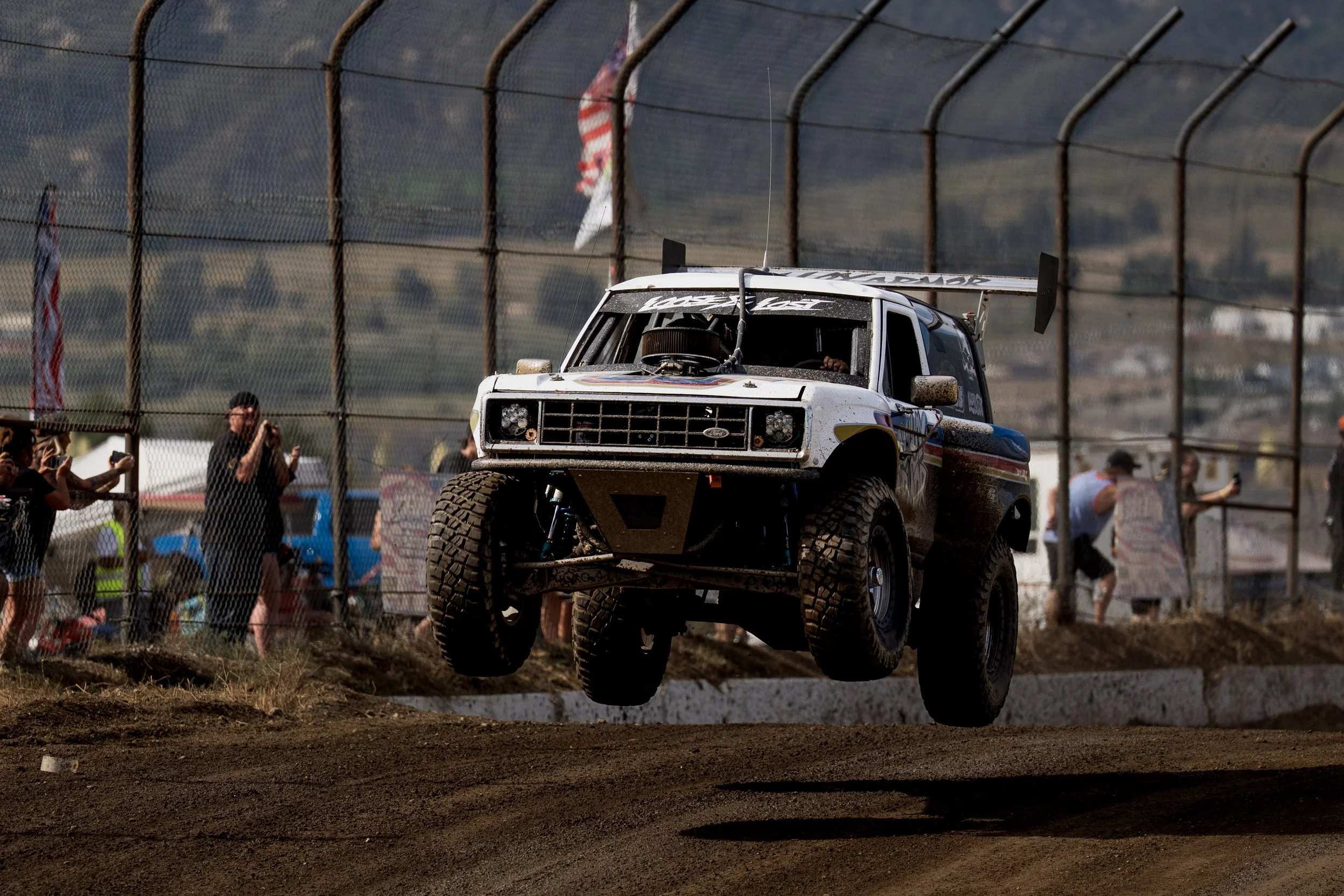 A racing truck airborne above dirt track during a race, with spectators and American flags in the background.