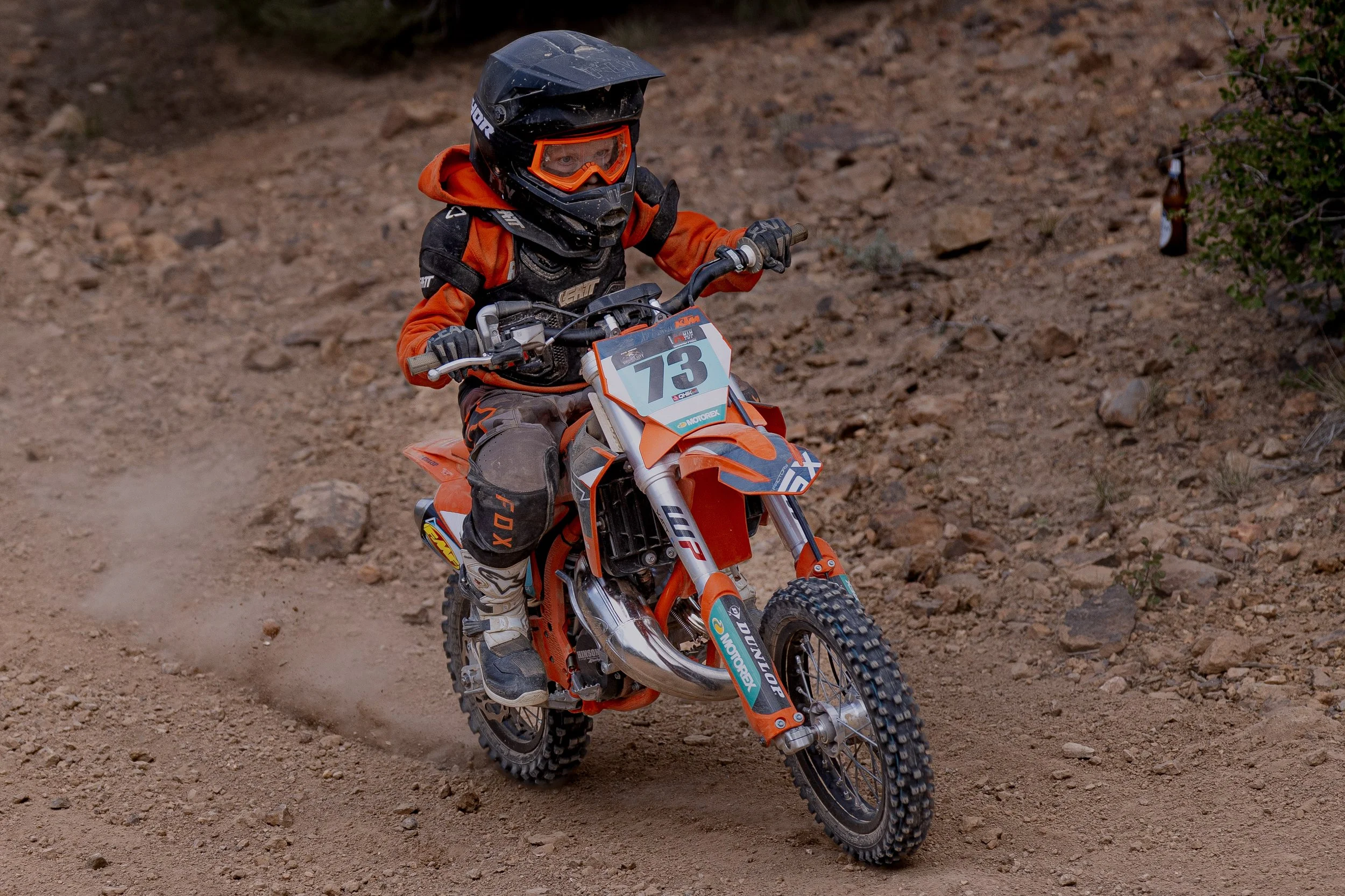 A young child wearing a full-face helmet and orange riding gear riding a KTM dirt bike on a dirt trail.