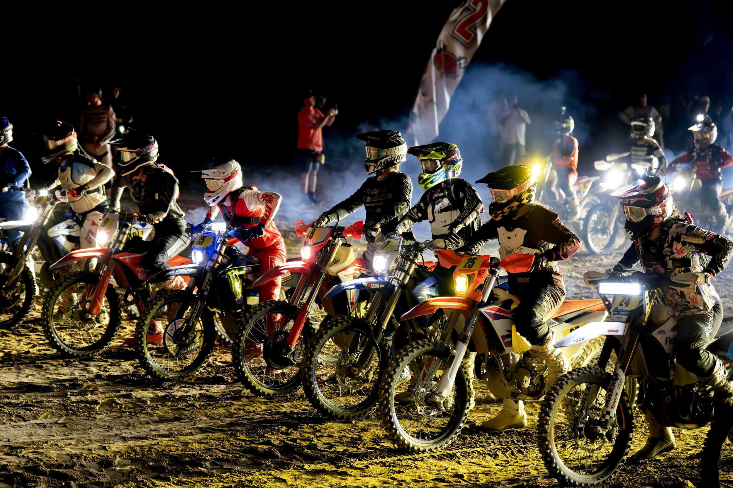 Motocross racers lined up on a dirt track at night, illuminated by their bike lights and some with helmets on, with spectators and flags in the background.