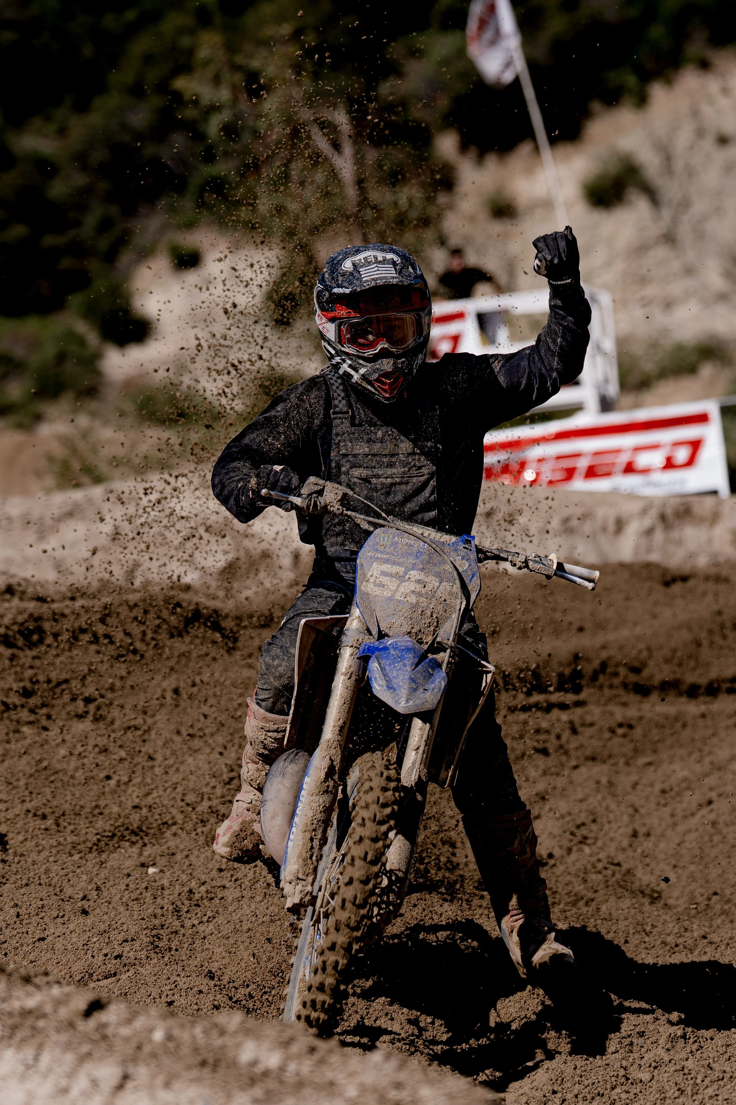 A motocross rider on a dirt track, wearing a helmet, goggles, and black gear, waving with mud flying around as they ride