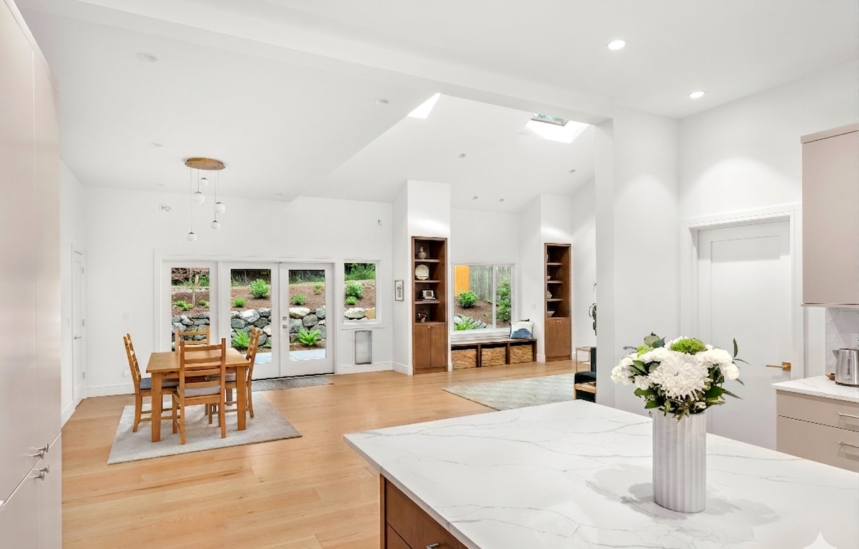 Open-concept kitchen and dining area addition project with white walls, wooden flooring, a dining table with four chairs, built-in cabinetry, and sliding glass doors opening to a garden with rocks and greenery.