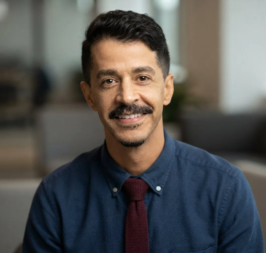 Headshot of a man with dark hair, a mustache, and a goatee, wearing a dark blue shirt and maroon tie, smiling in a professional setting.