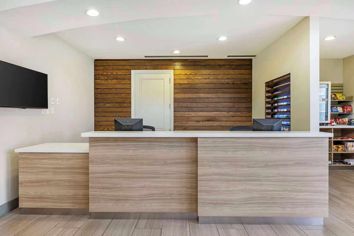 Empty hotel or reception desk with two computer monitors, a wooden accent wall behind, and snack shelves on the side.
