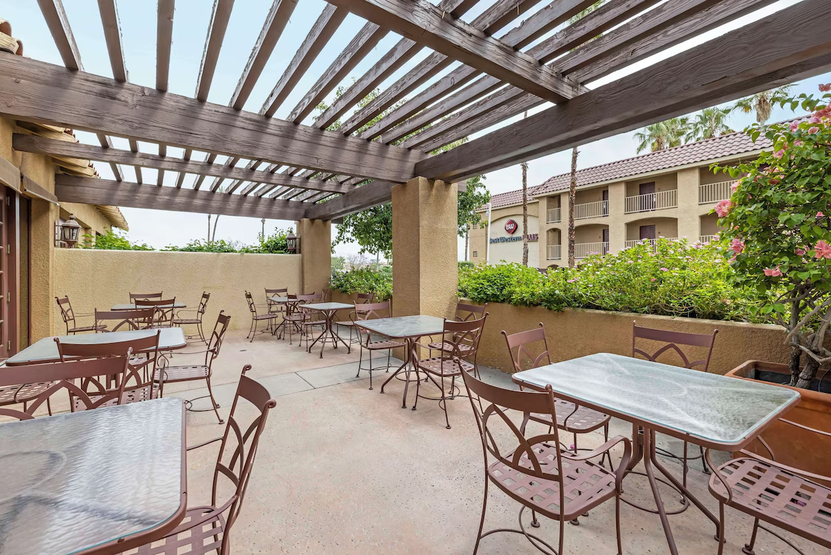Outdoor patio with metal tables and chairs under a wooden pergola, surrounded by greenery and nearby buildings.