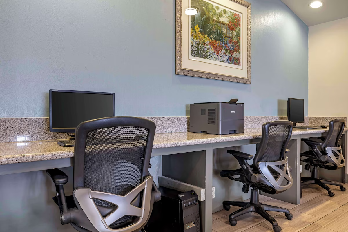 Workstations with computers, office chairs, a printer, and framed artwork on a wall in an office or business center