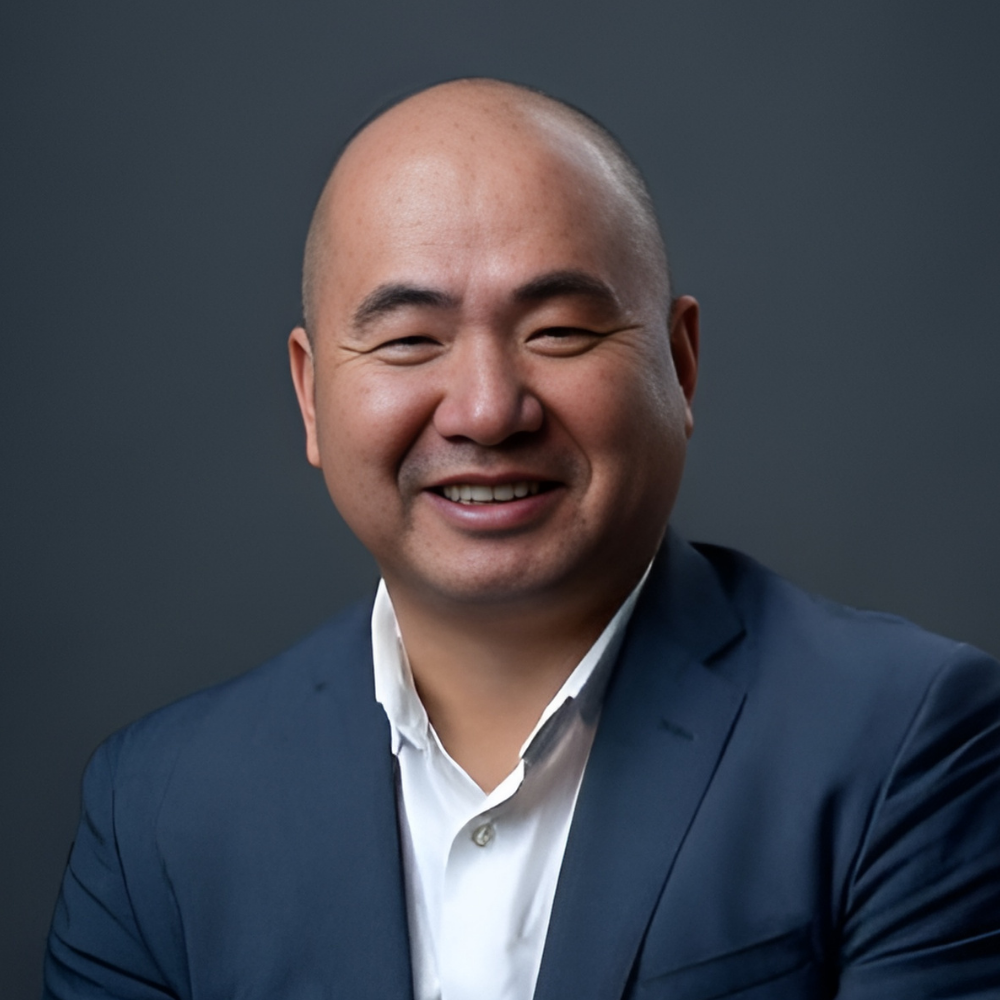 A man with a bald head, smiling, wearing a dark suit jacket over a white shirt, posing in front of a dark background.